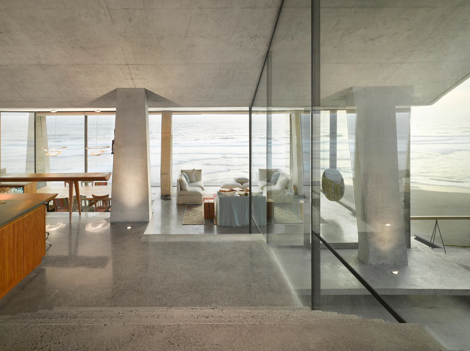 A minimalist coastal living room featuring raw concrete walls, polished floors, structural pillars, and a glass partition overlooking the ocean.