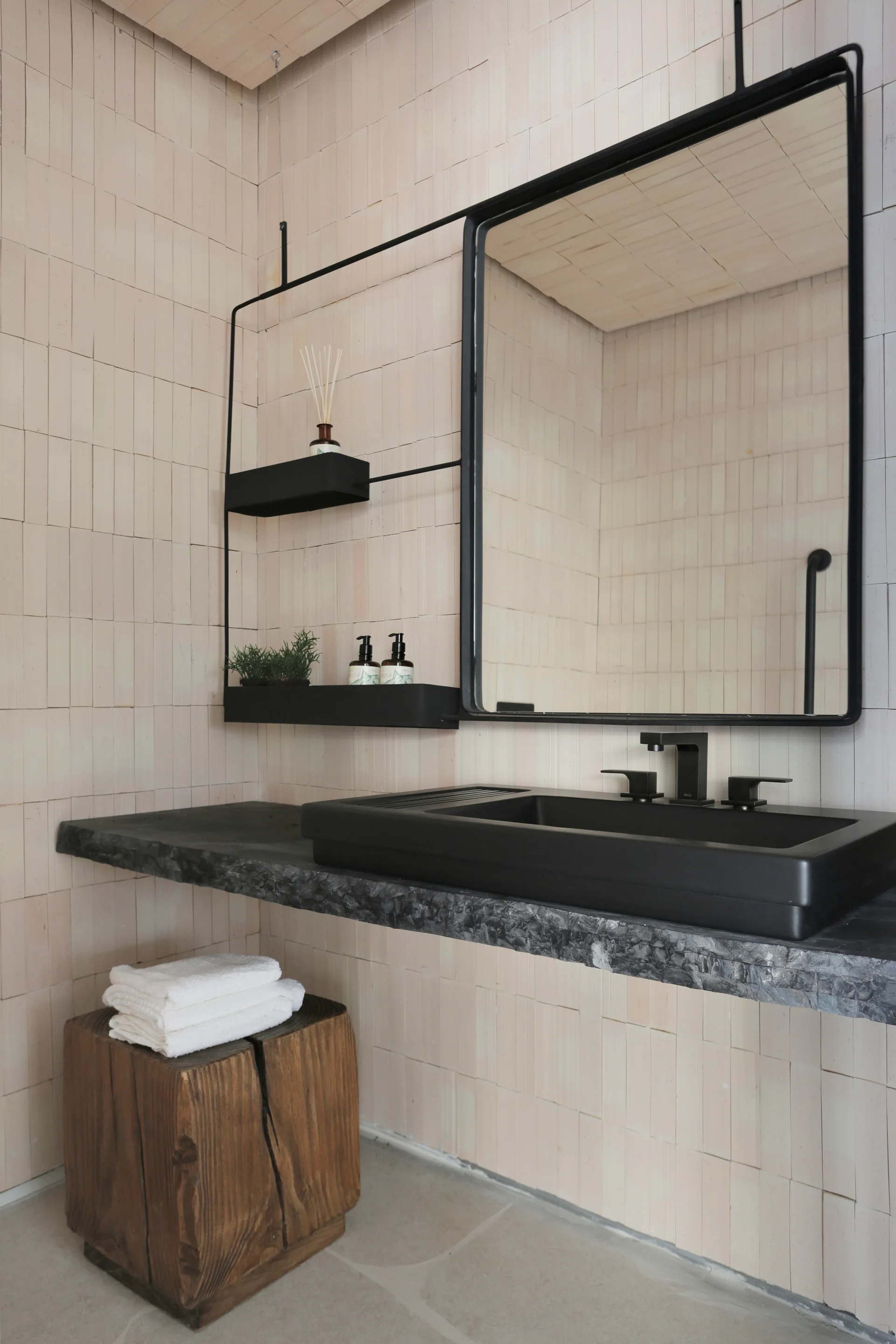 A modern bathroom vanity features a matte black sink on a dark stone slab, a rectangular mirror, and a wall clad in vertical beige tiles.