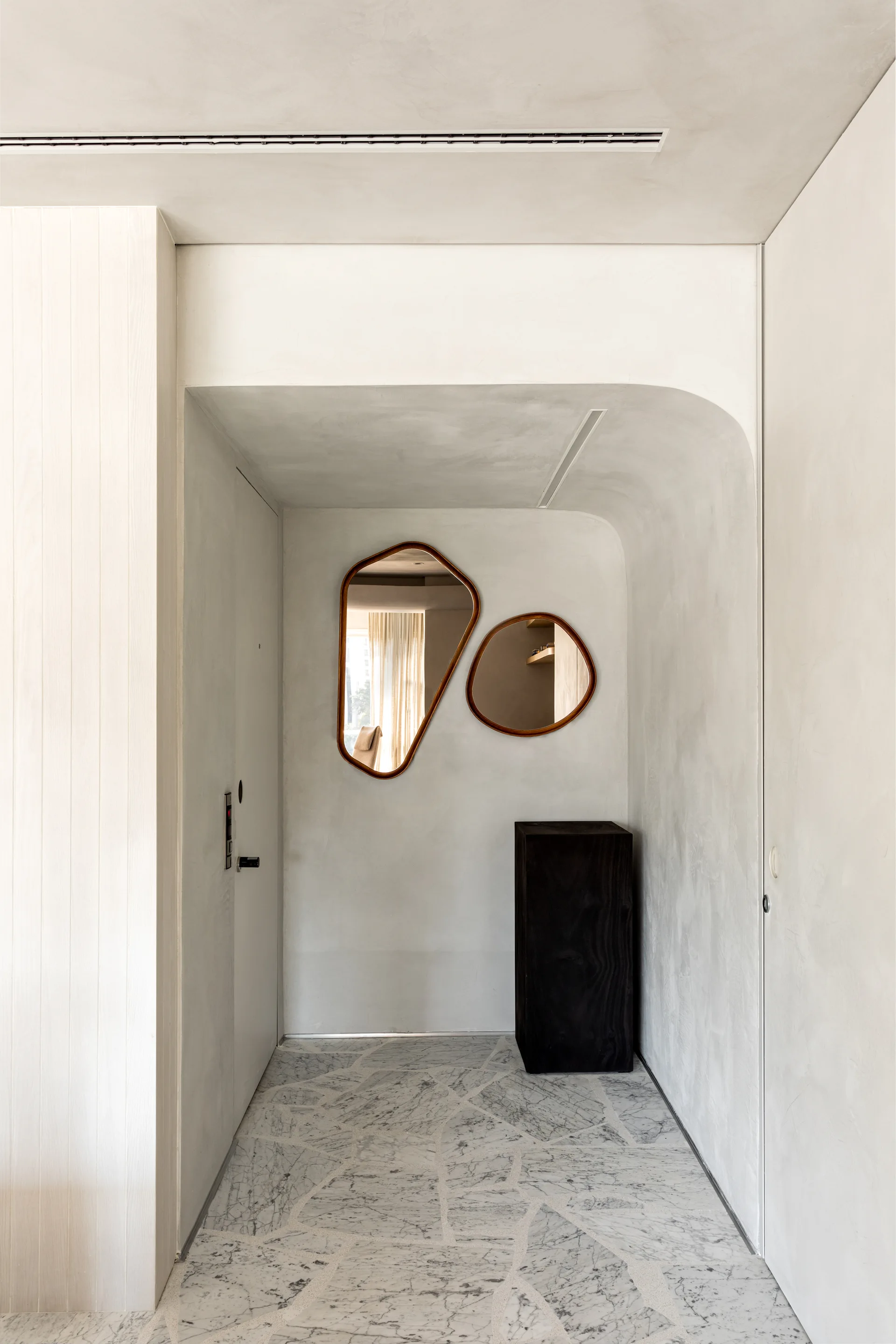 A minimalist entryway features a curved plaster wall with two organic-shaped wooden mirrors, a dark wooden pedestal, and an irregular stone tile floor.