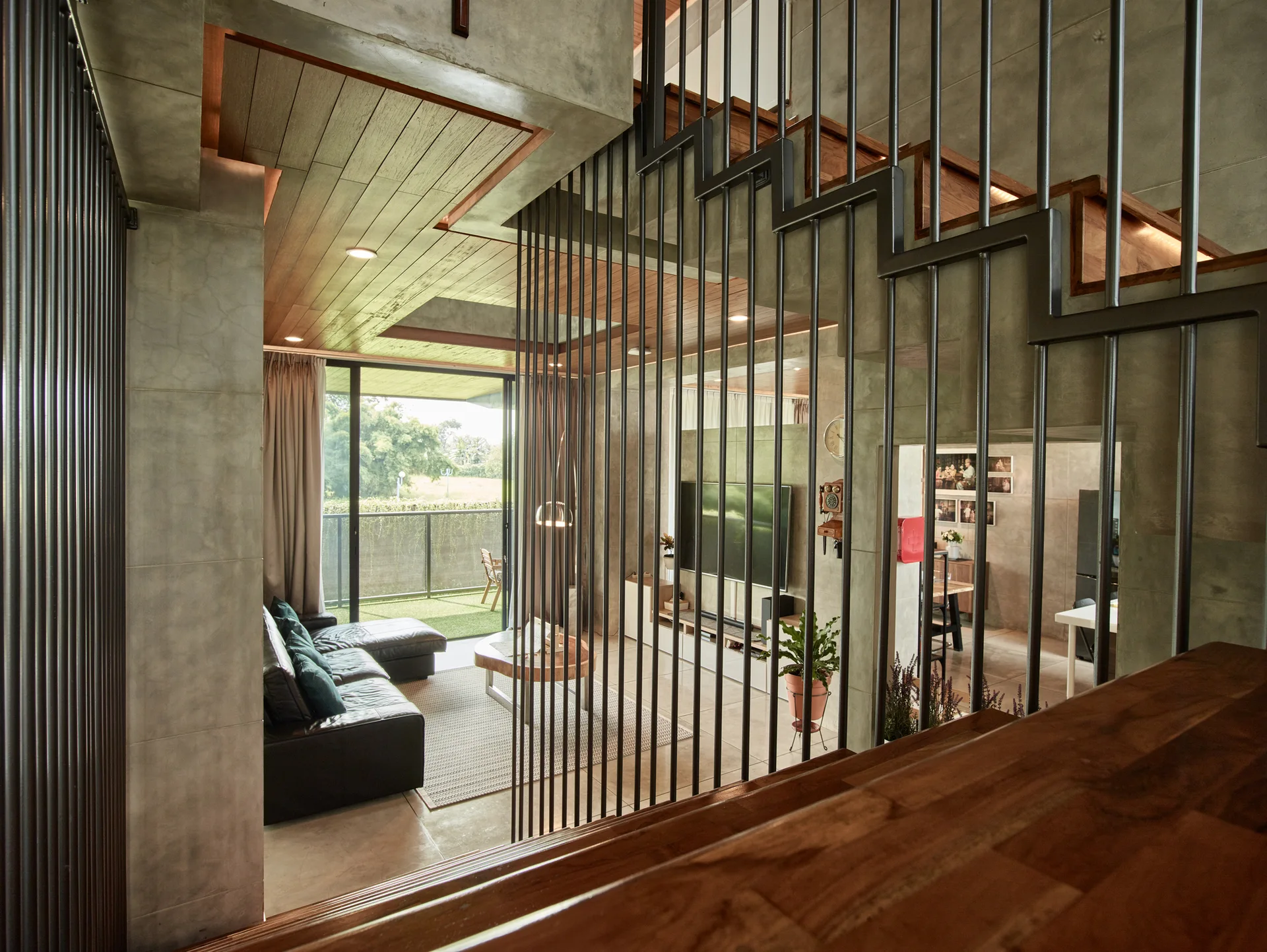 A modern living room with a black sectional and concrete walls viewed through a vertical black metal rod partition next to wooden stairs.