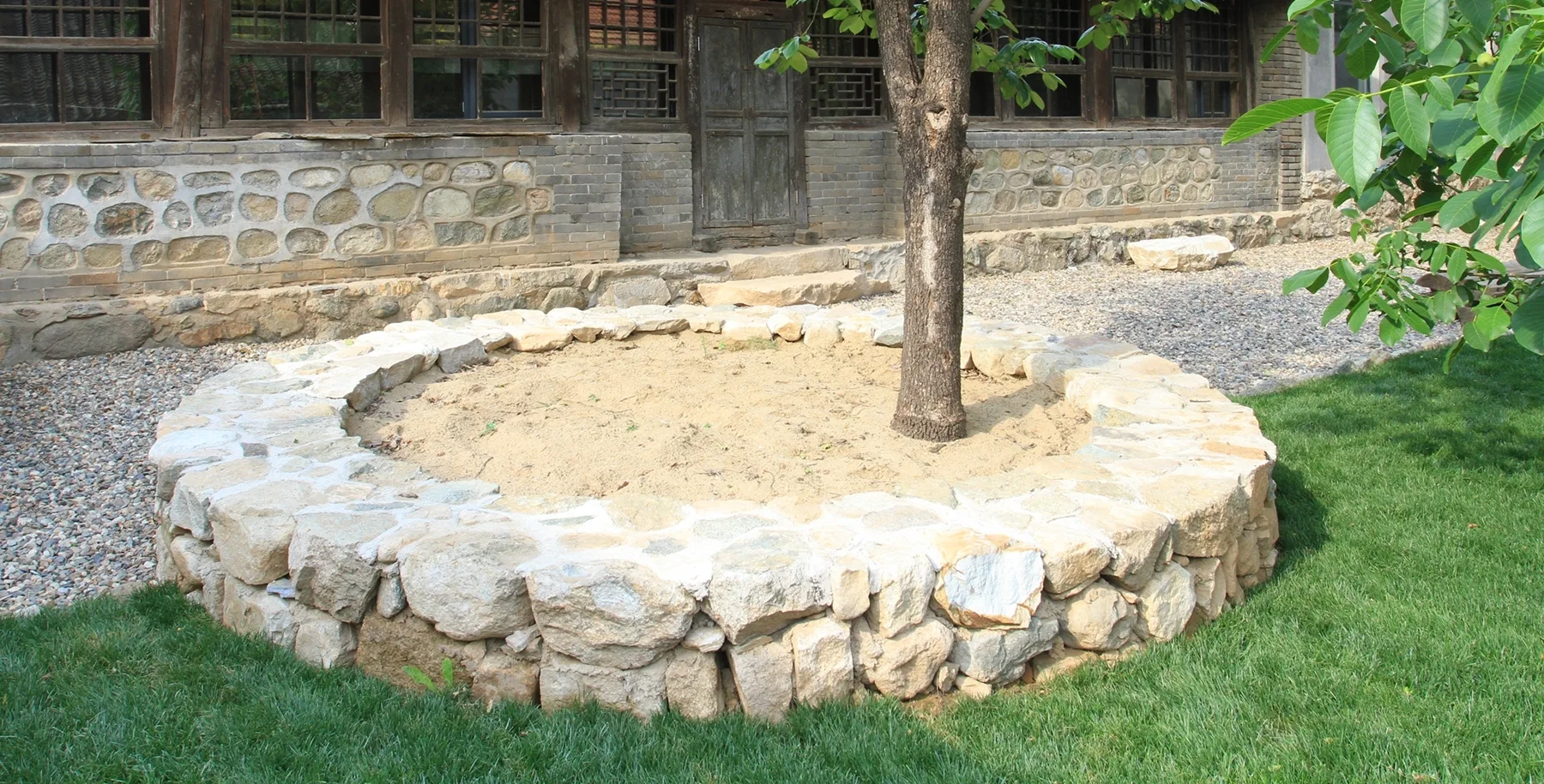 A circular raised stone planter surrounds a tree trunk in a garden featuring a green lawn, gravel path, and a rustic stone building in the background.