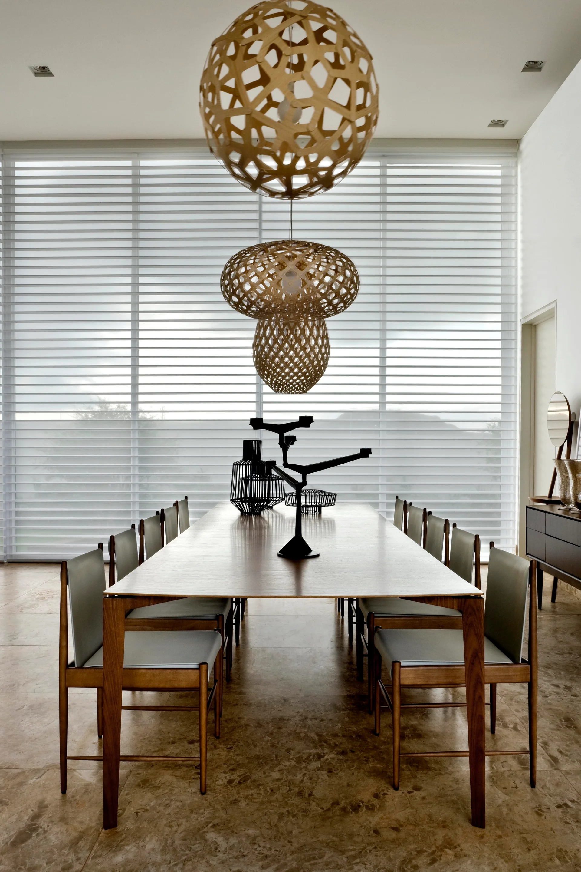 A long wooden dining table and matching chairs sit under three vertically stacked woven pendant lights in front of a window with white horizontal blinds.