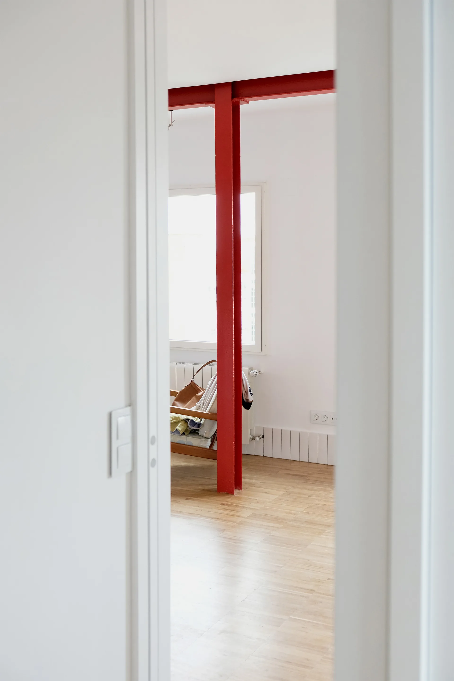 A view through a white doorway into a minimalist room with a bright red steel column, light wood floors, and a window.