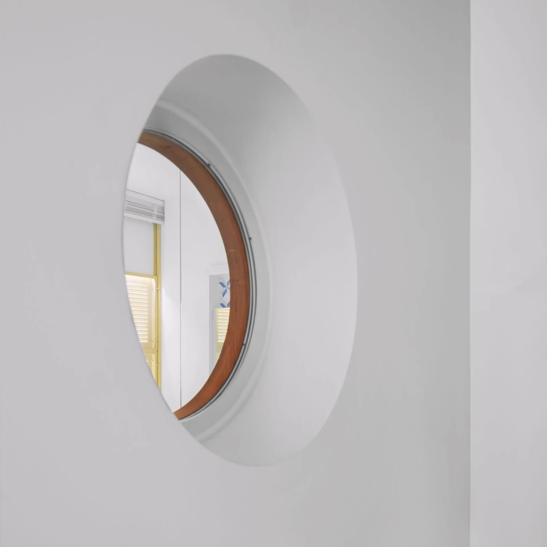 A smooth white wall features a circular window opening with a warm brown wood frame, showing a yellow shutter inside.