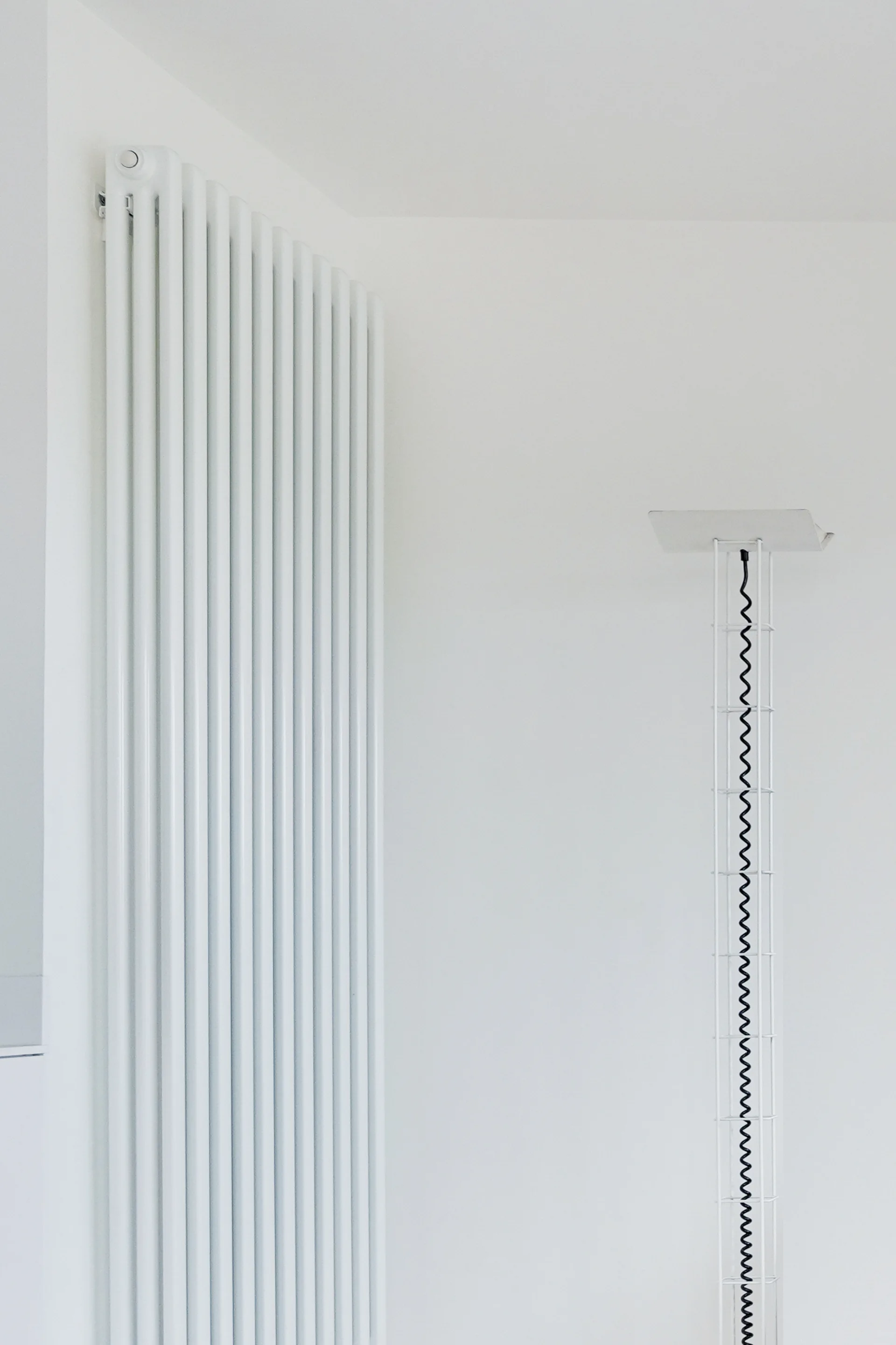 A tall white vertical radiator and a slender white floor lamp with a black spiral cord stand against a plain white wall.
