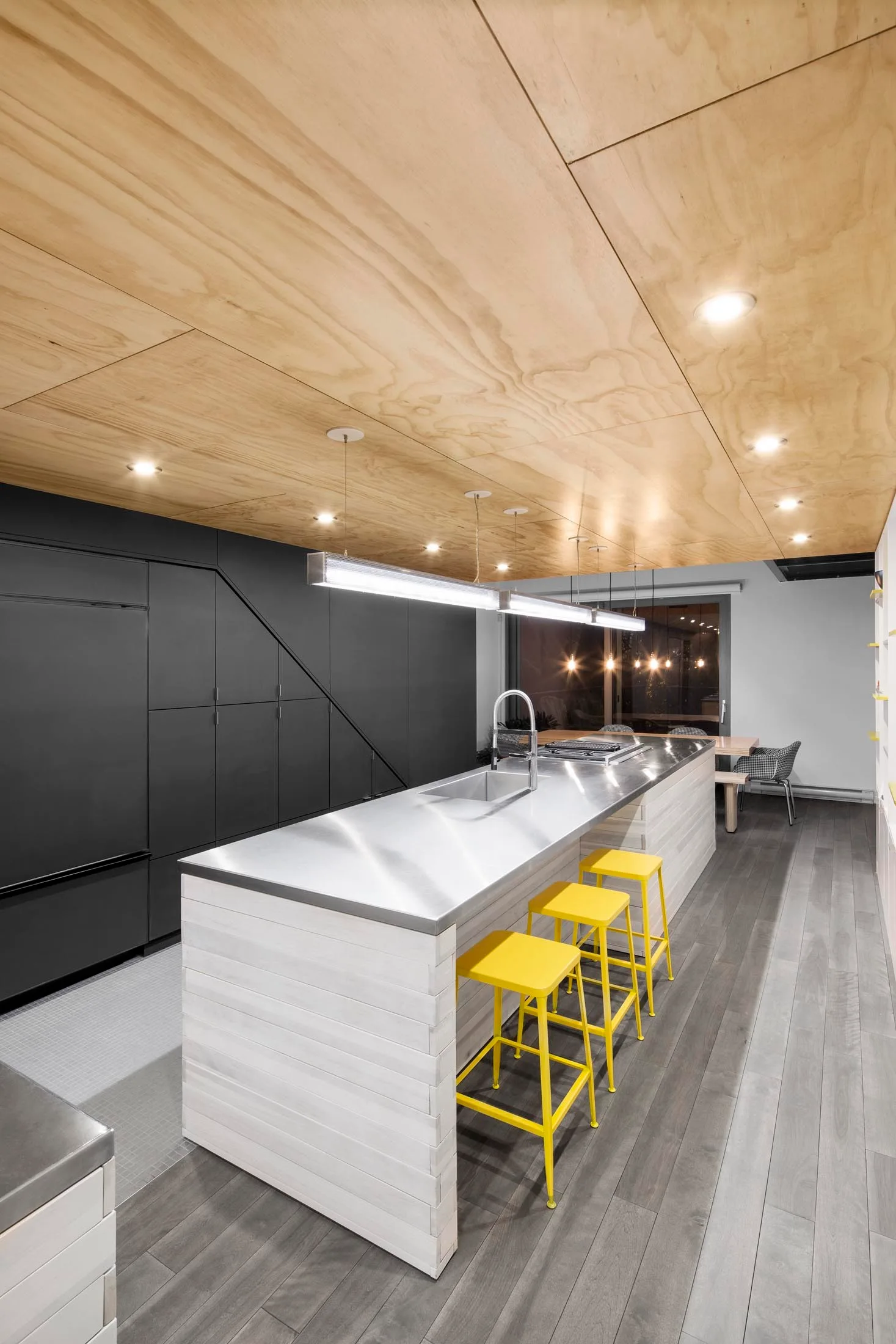 A modern kitchen featuring a stainless steel island with three yellow stools, a plywood paneled ceiling, and dark matte cabinetry.