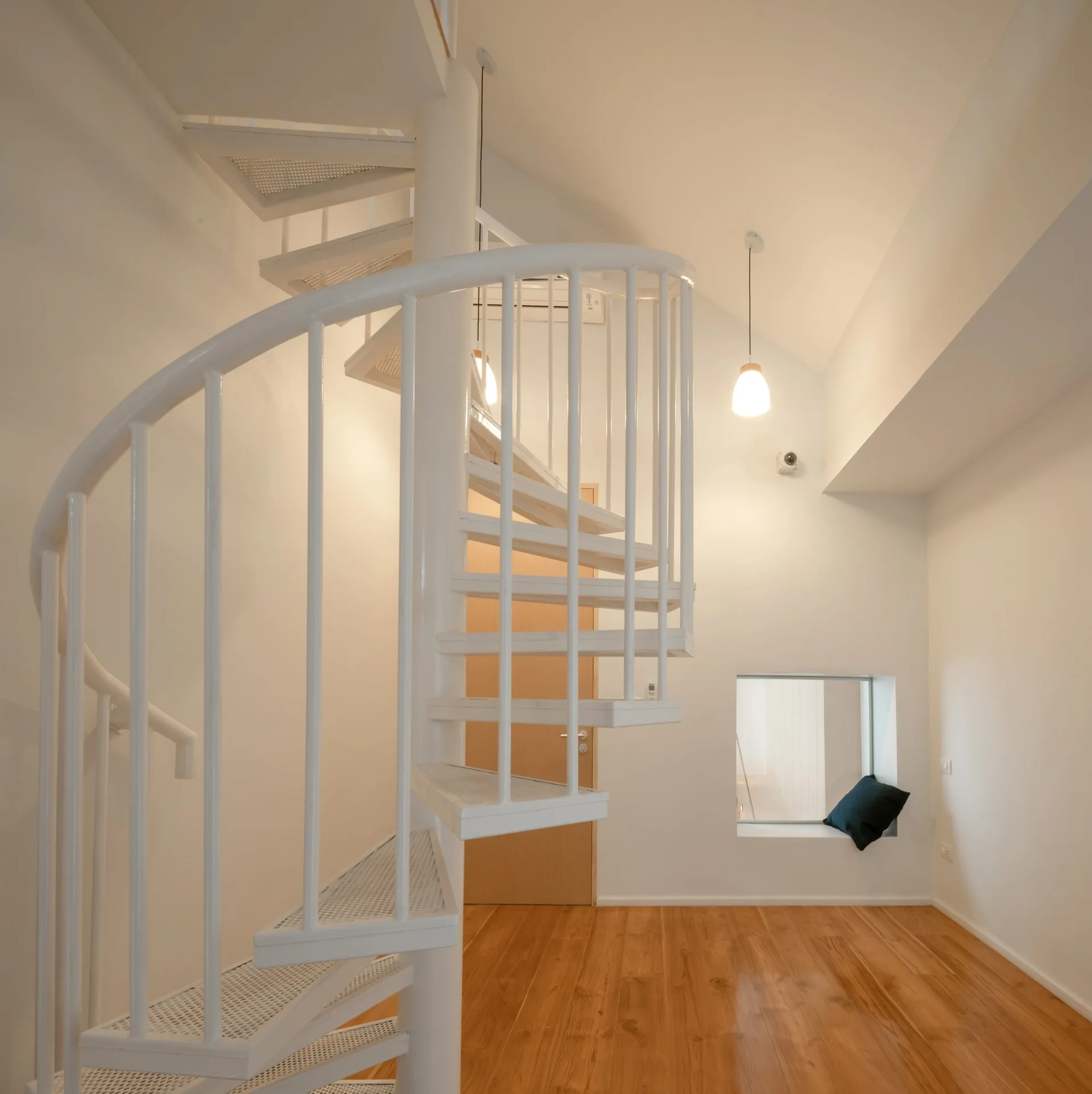 A white metal spiral staircase stands on polished hardwood flooring in a minimalist room with a recessed square window nook and a sloped ceiling.