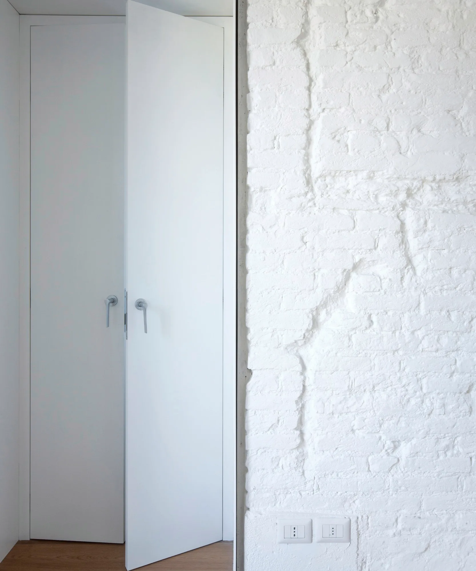 Minimalist matte white closet doors with slim metal handles stand next to a rough, white-painted brick wall.