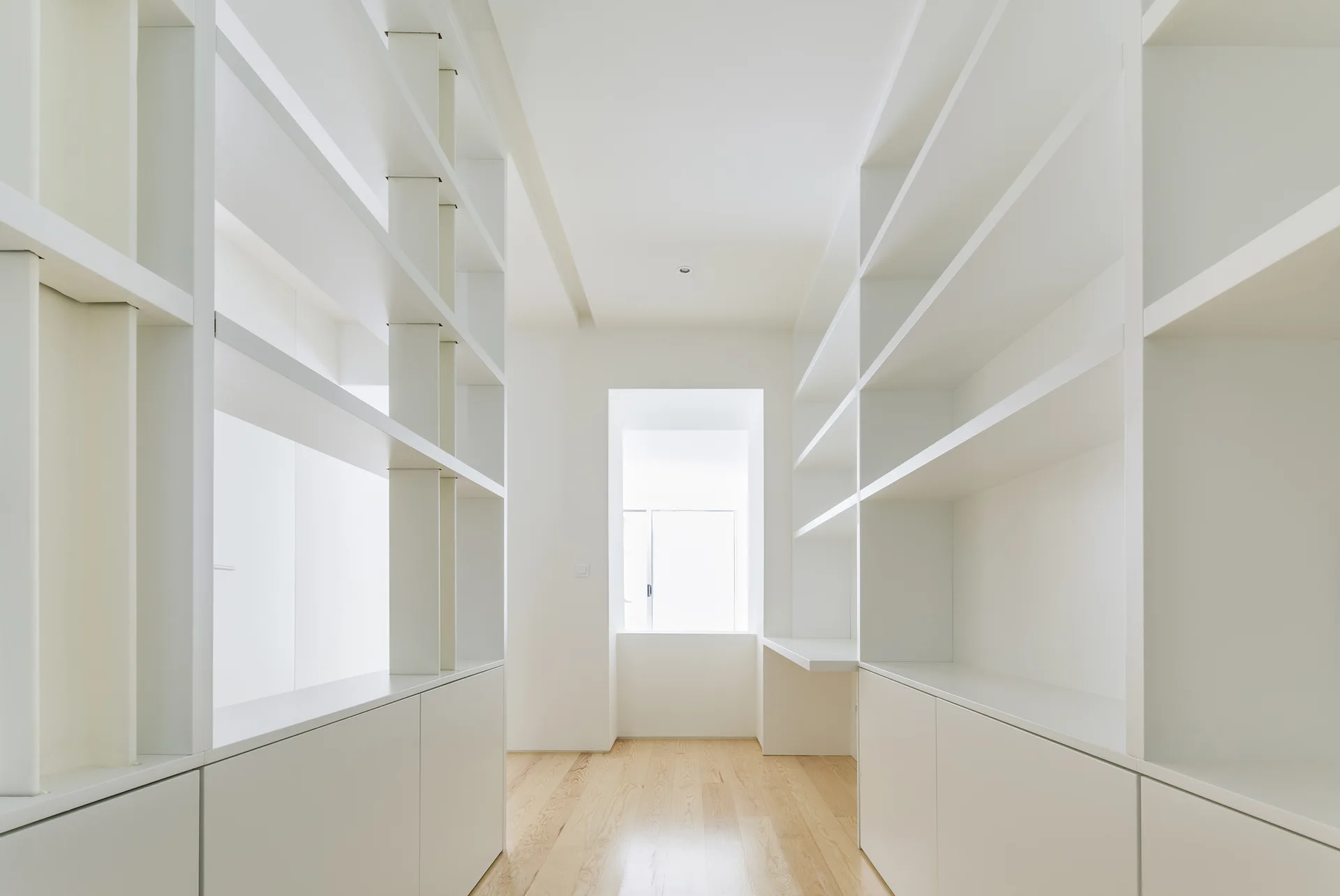 A bright white hallway features symmetrical floor-to-ceiling built-in shelving units and natural wood flooring leading toward a window.