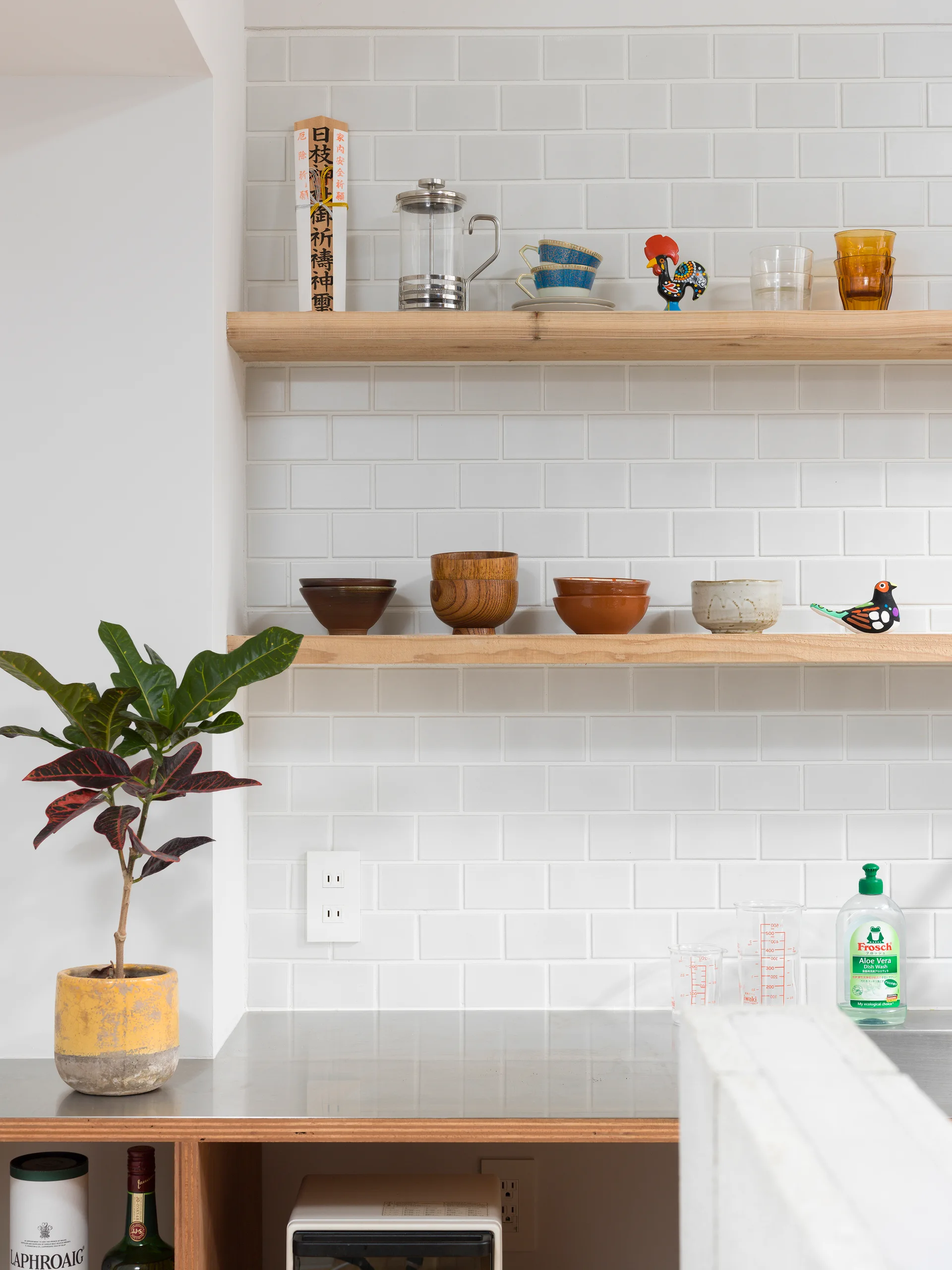 Two light wood open shelves are mounted on a white subway tile wall above a metal kitchen counter with a yellow potted plant.