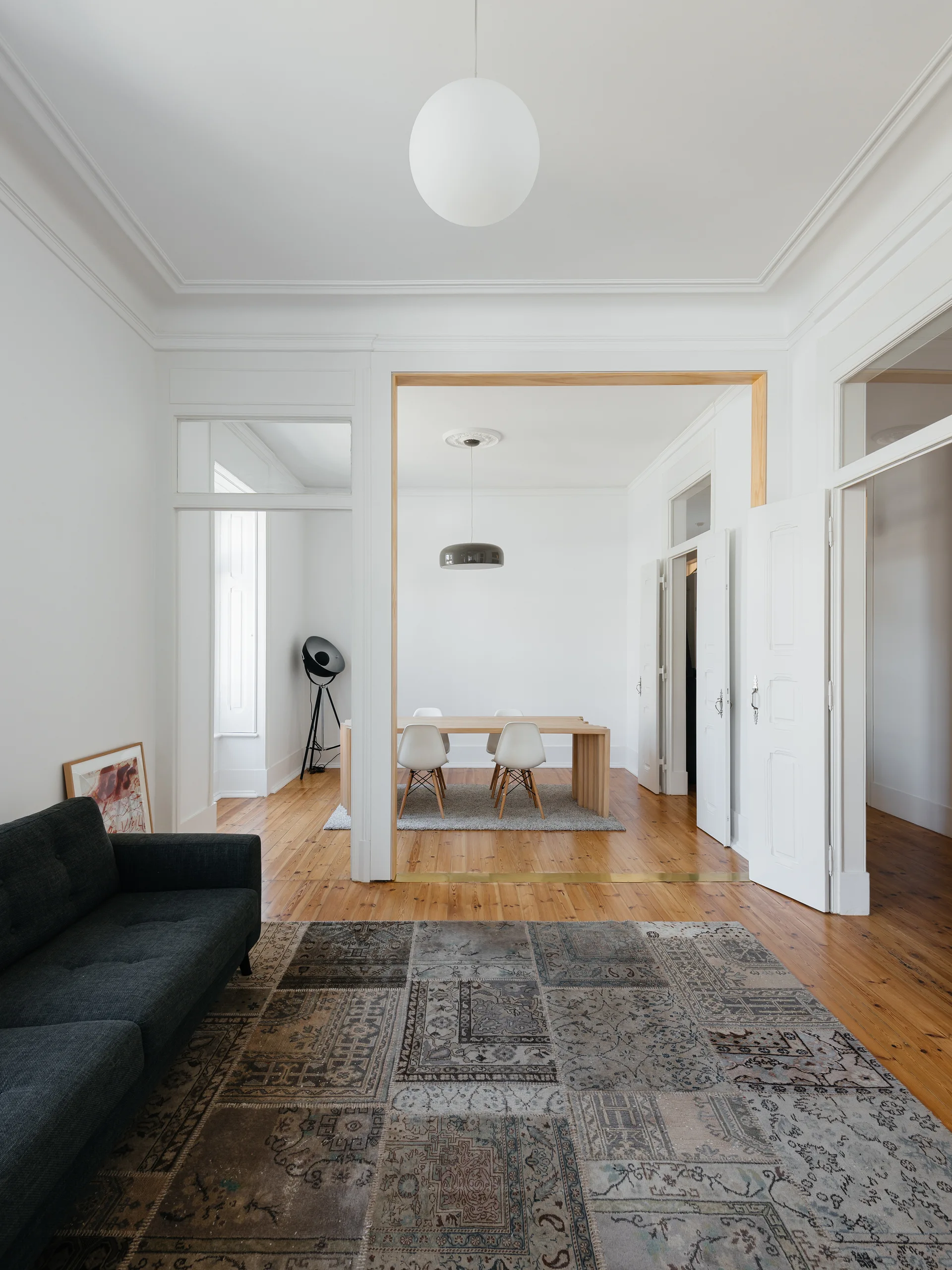 A bright living room with a dark grey sofa and patchwork rug leads through a wide wooden archway to a minimalist dining room.