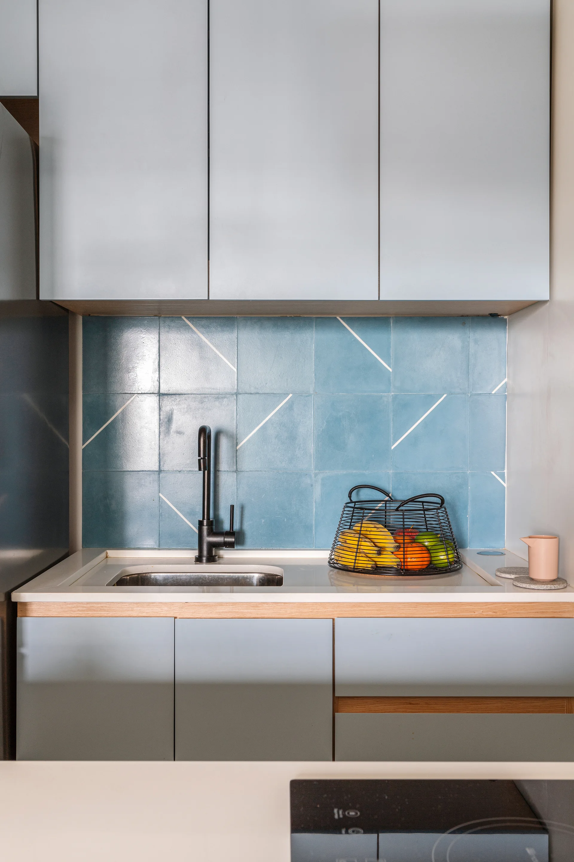 A modern kitchen sink area features light blue cabinets, a blue square-tiled backsplash with diagonal lines, and a black gooseneck faucet.