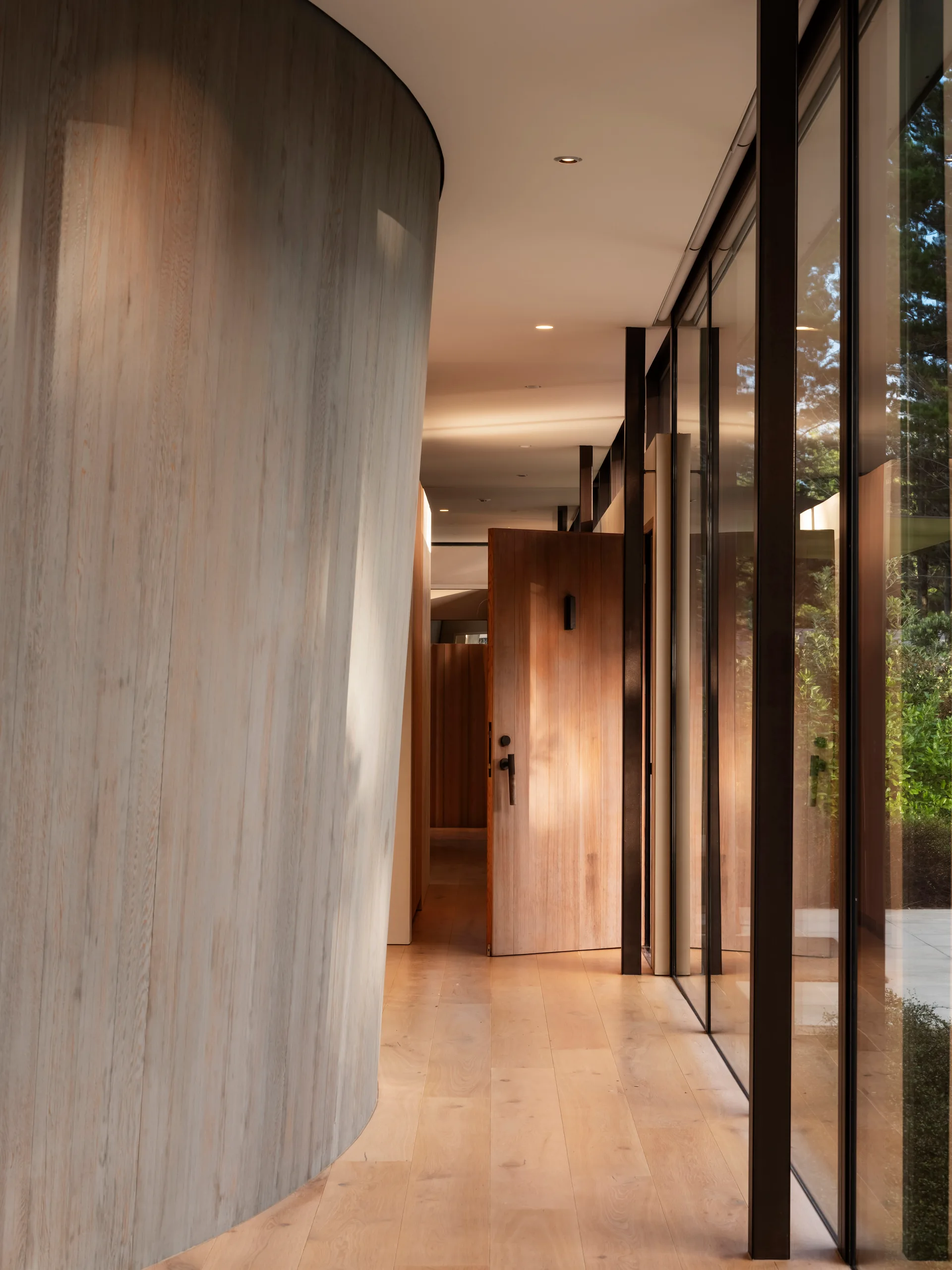 A minimalist hallway featuring a prominent curved wood-clad wall and floor-to-ceiling glass windows.