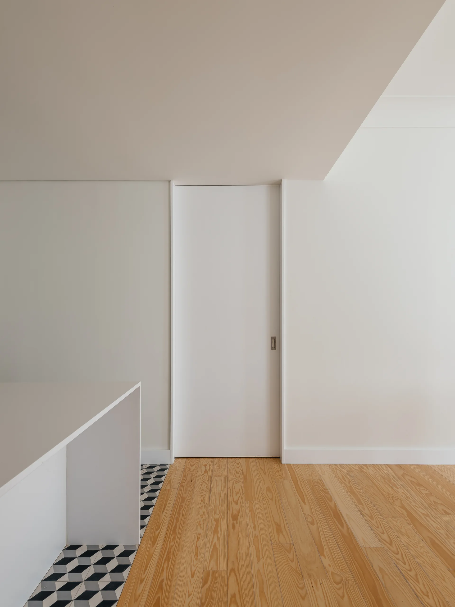 A white sliding pocket door stands in a minimalist hallway featuring a transition from natural wood floor planks to black and white geometric floor tile.