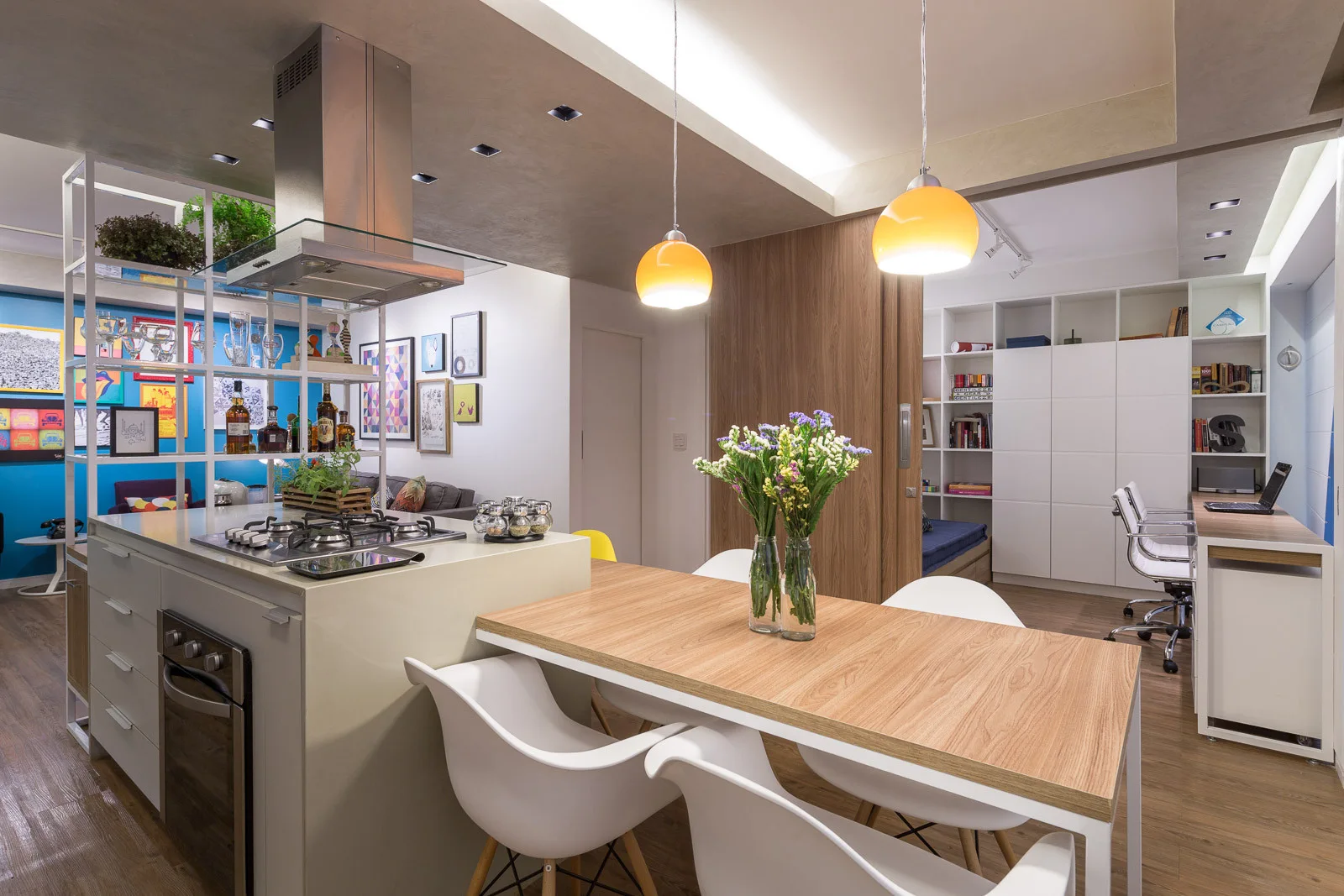 A kitchen island with an attached wood dining table sits under orange pendant lights next to a workspace with white built-in shelving.