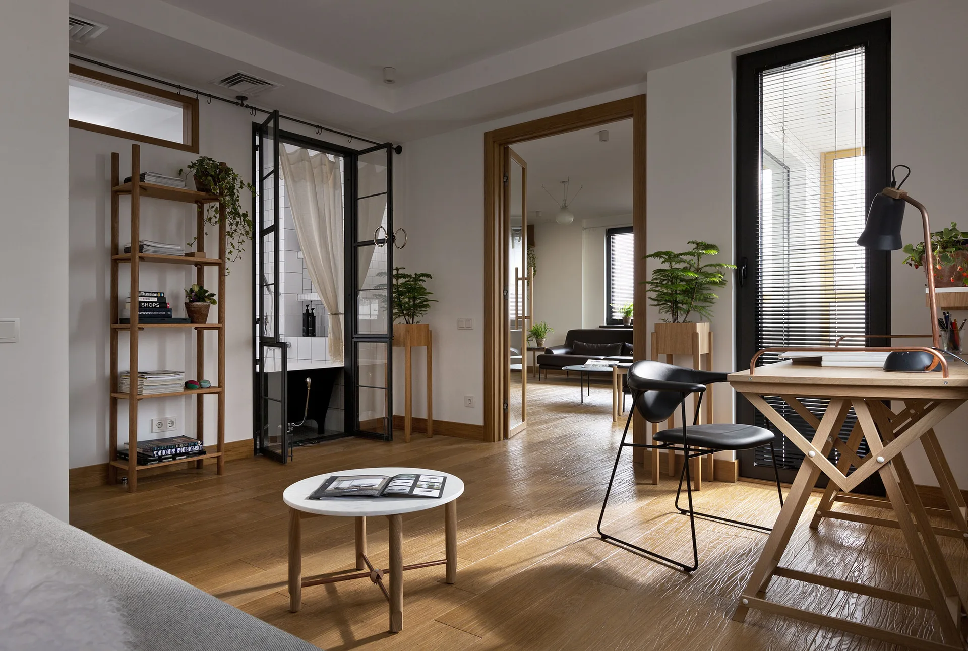 A bright modern living room with light oak flooring, a wooden workspace desk with black chair, and a black-framed glass door opening to a bathroom.