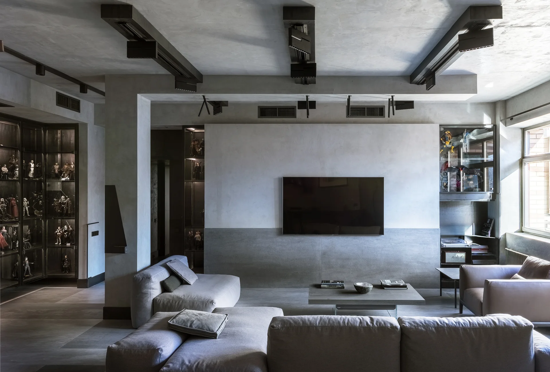 A minimalist grey living room featuring a low sectional sofa, textured concrete walls, ceiling beams, and integrated dark glass display cabinets.