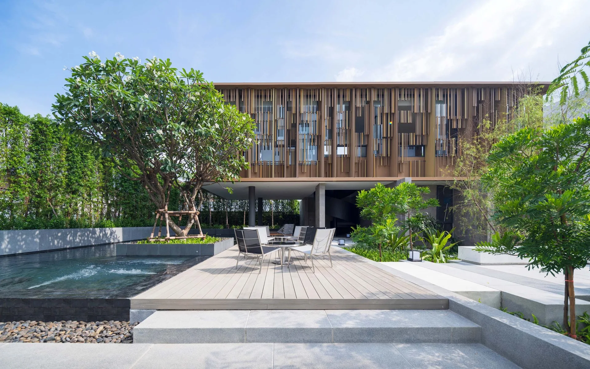 A modern outdoor patio features a wooden deck with seating next to a pool and a contemporary building with a louvered wood facade.