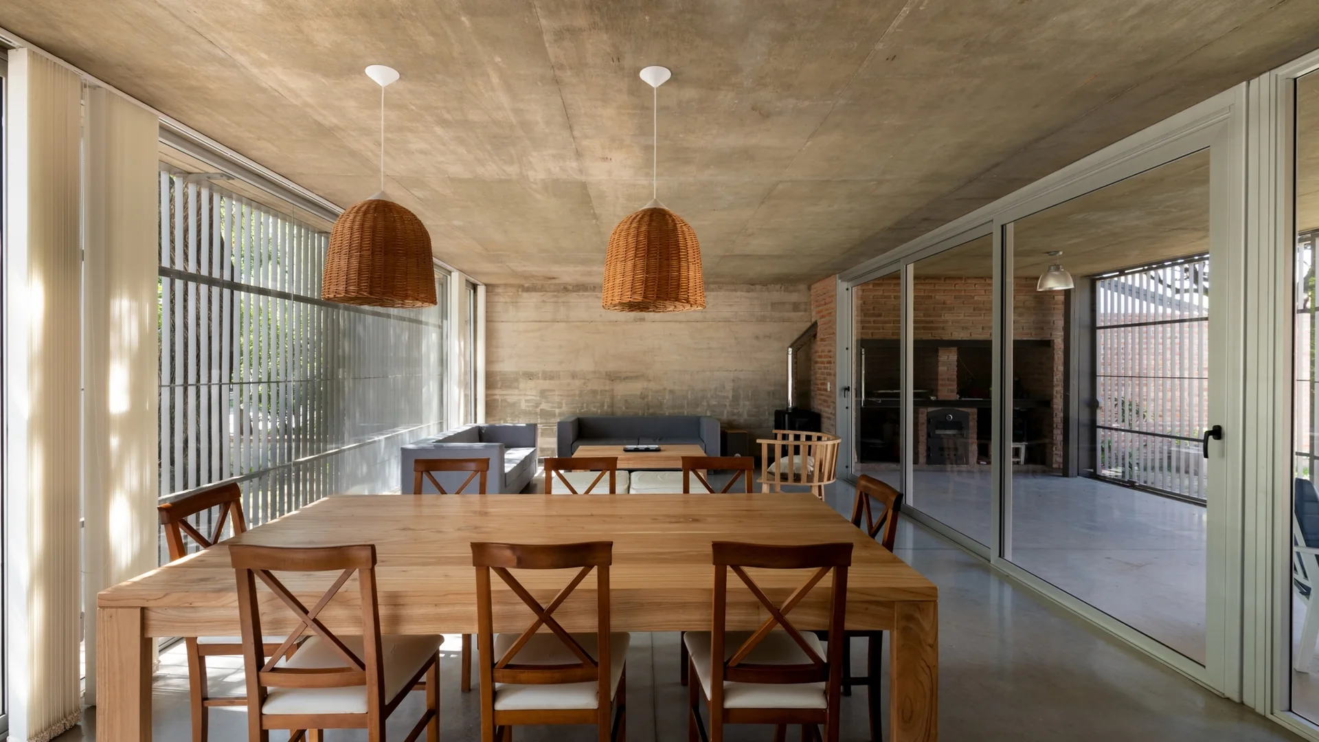 An open-plan dining area features a large wooden dining table with chairs, wicker pendant lights, and raw concrete ceilings and floors.