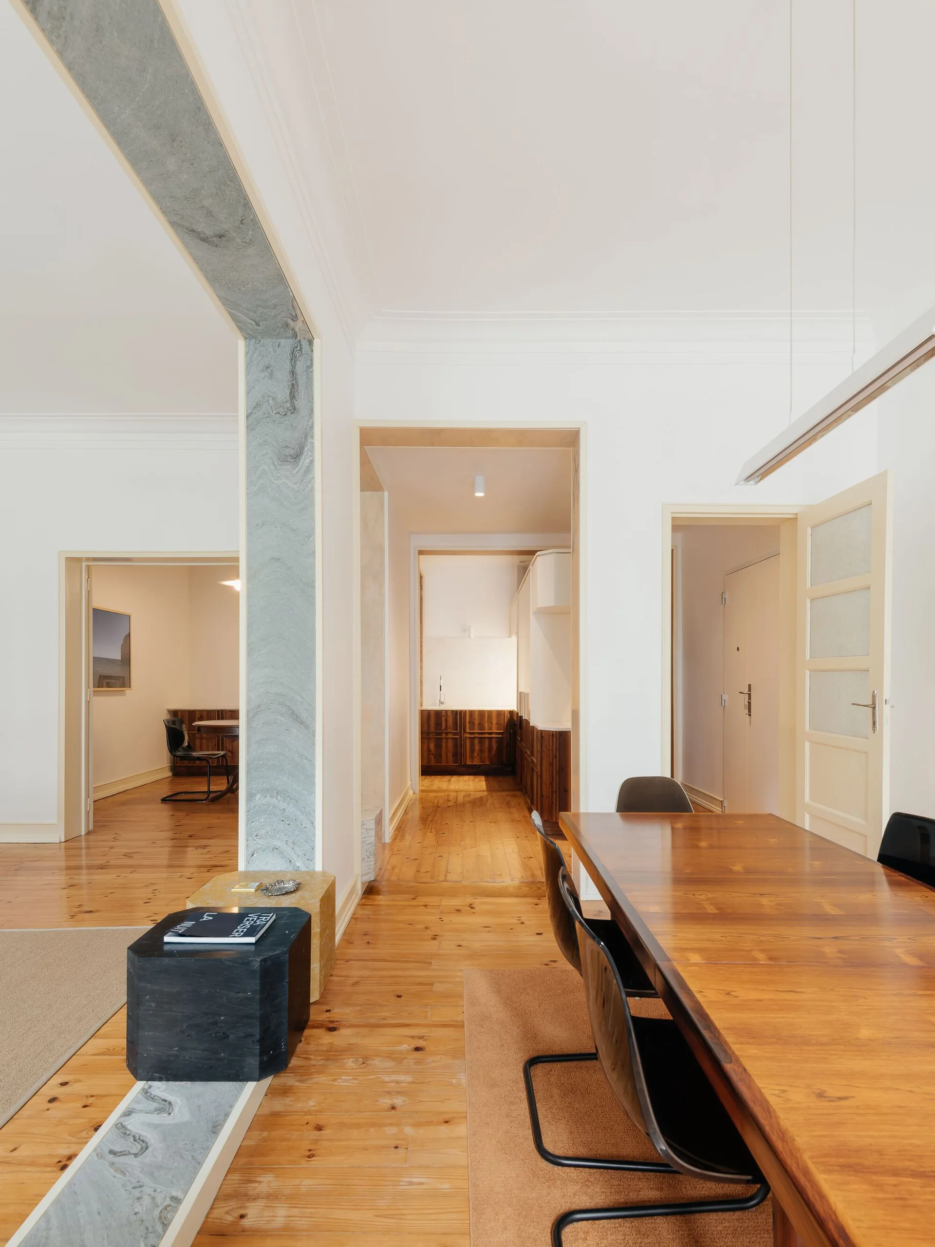 A wooden dining table with black chairs sits on wood floors next to a gray veined stone archway leading to adjoining rooms.