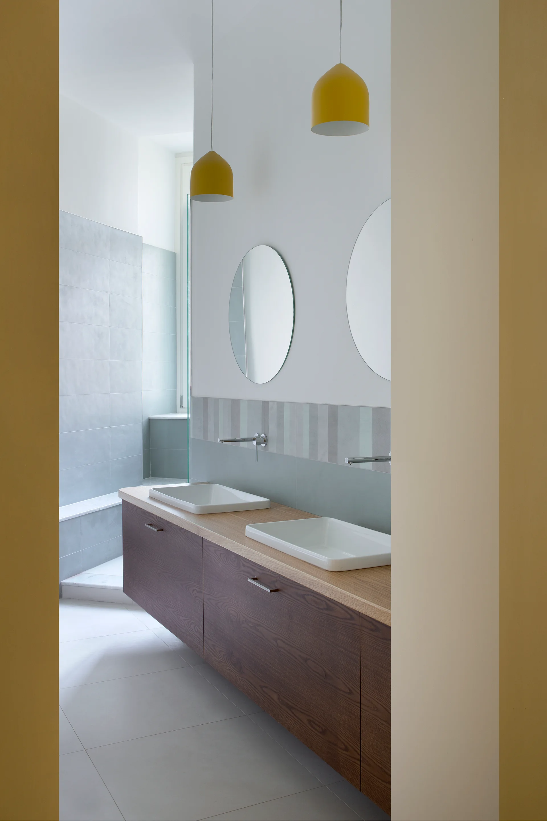 A modern bathroom viewed through a doorway, showing a floating wooden double vanity, two round mirrors, and two yellow pendant lights.