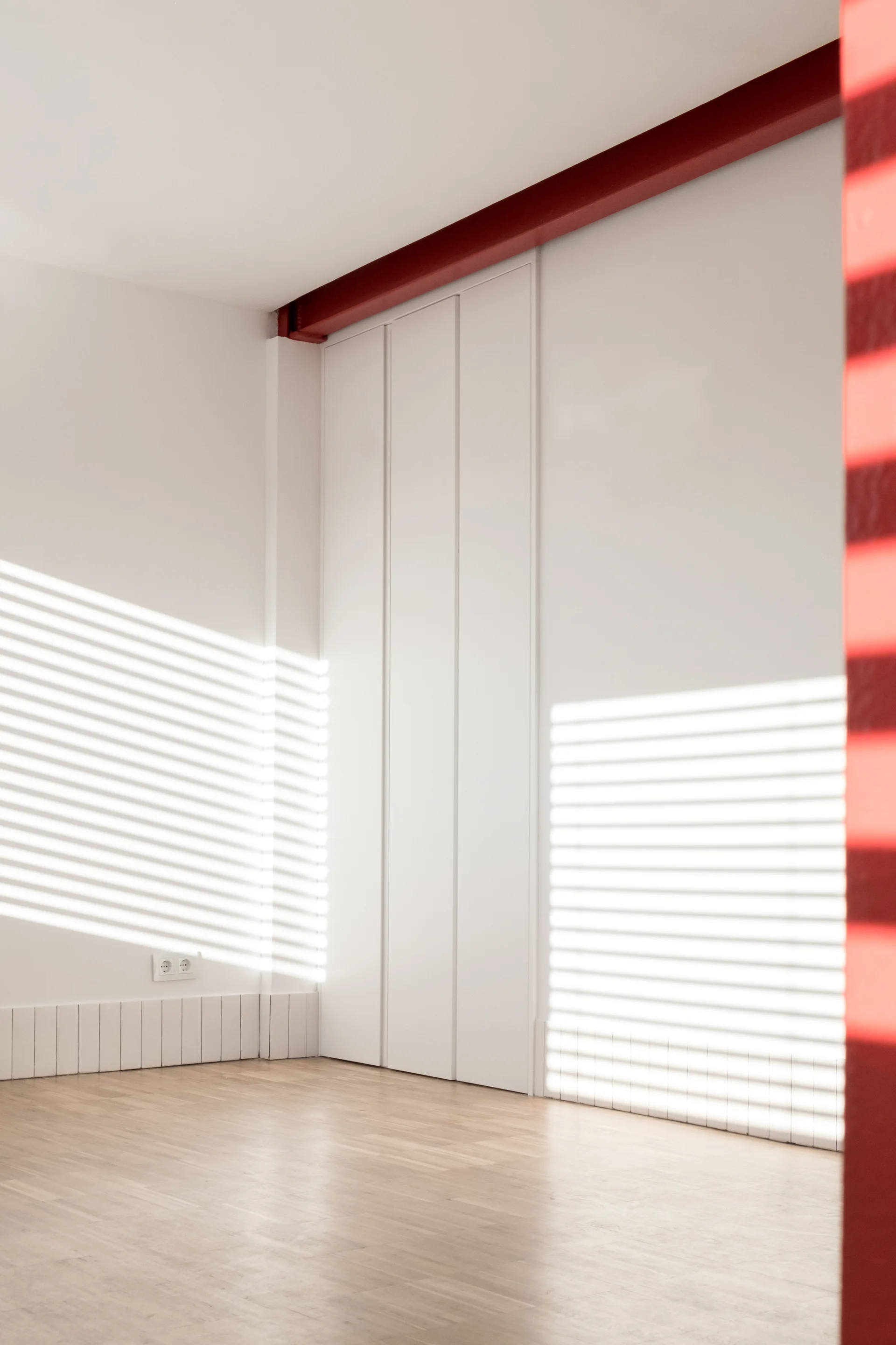 An empty minimalist room featuring a red painted ceiling beam, white walls, vertical closet panels, and striped light patterns on the wood floor.