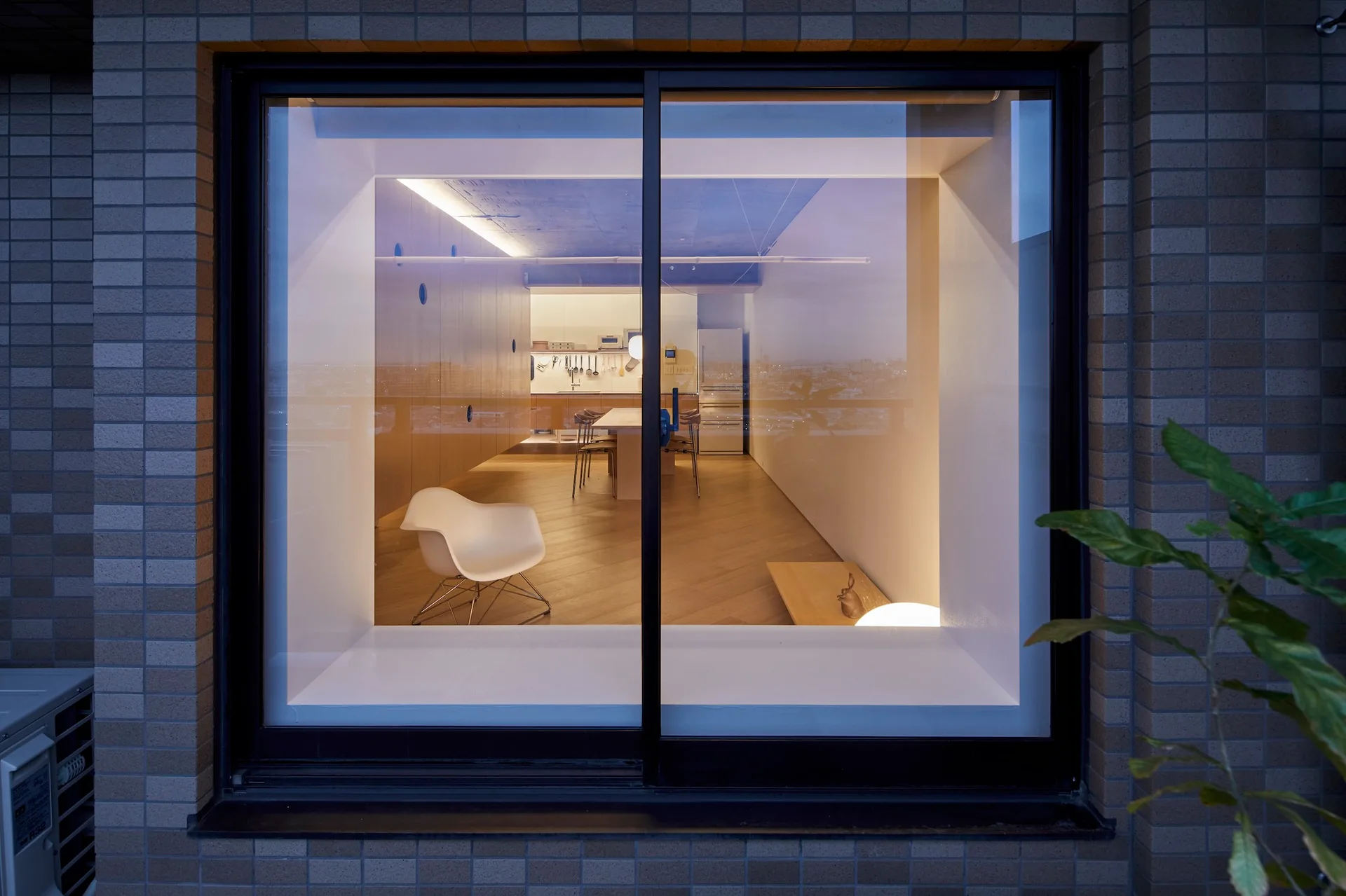 A view through a large black-framed window into a minimalist kitchen and dining area with light wood floors and integrated cabinetry.