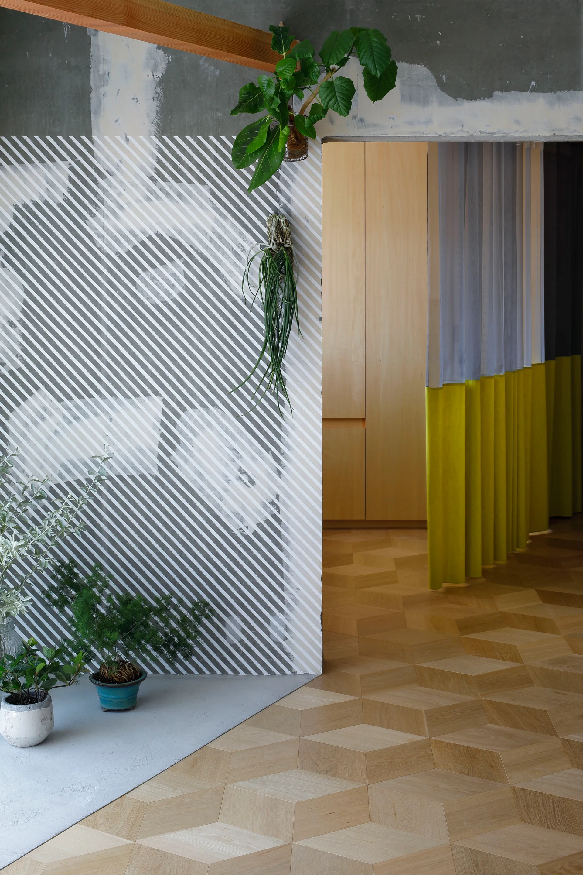 An interior view featuring a diagonal-striped wall partition, geometric parquet wood flooring, and a yellow color-blocked curtain in a doorway.