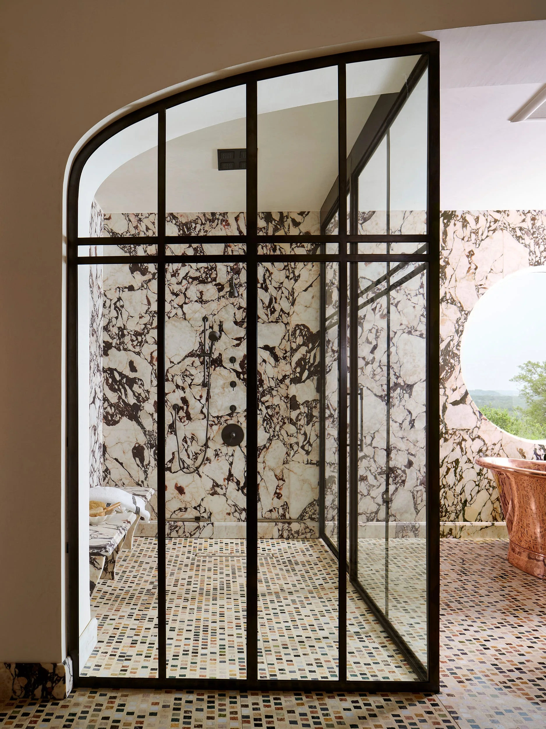 A bathroom featuring an arched walk-in shower with a black metal grid enclosure, heavily veined marble walls, and a copper tub near a round window.