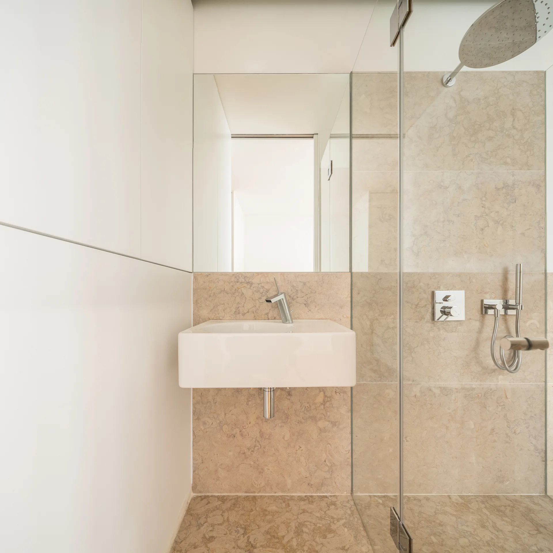 A minimalist bathroom features a white wall-mounted sink, a glass-enclosed shower, and beige stone tiles on the wall and floor.