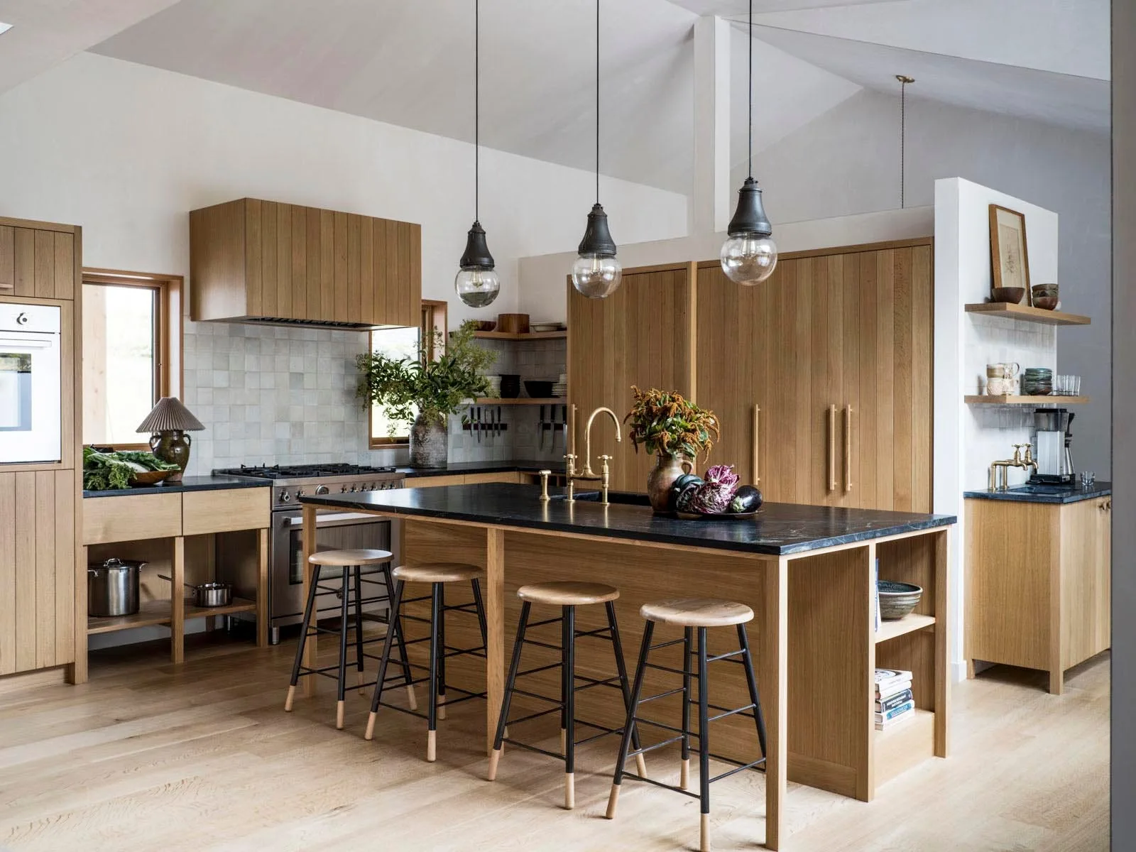 A light oak kitchen features a wood island with a dark stone countertop, four bar stools, three glass pendant lights, and a white tiled backsplash.
