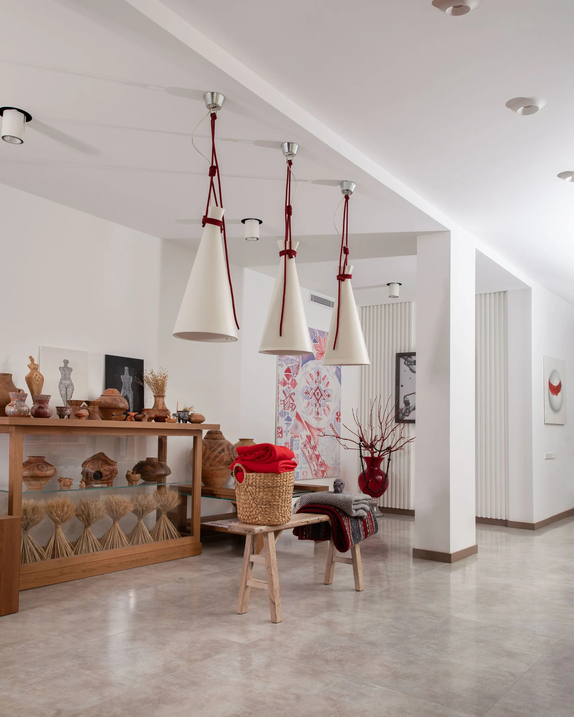A spacious white hallway featuring three white conical pendant lights hanging over a rustic wooden bench and a console table filled with ceramic pottery.