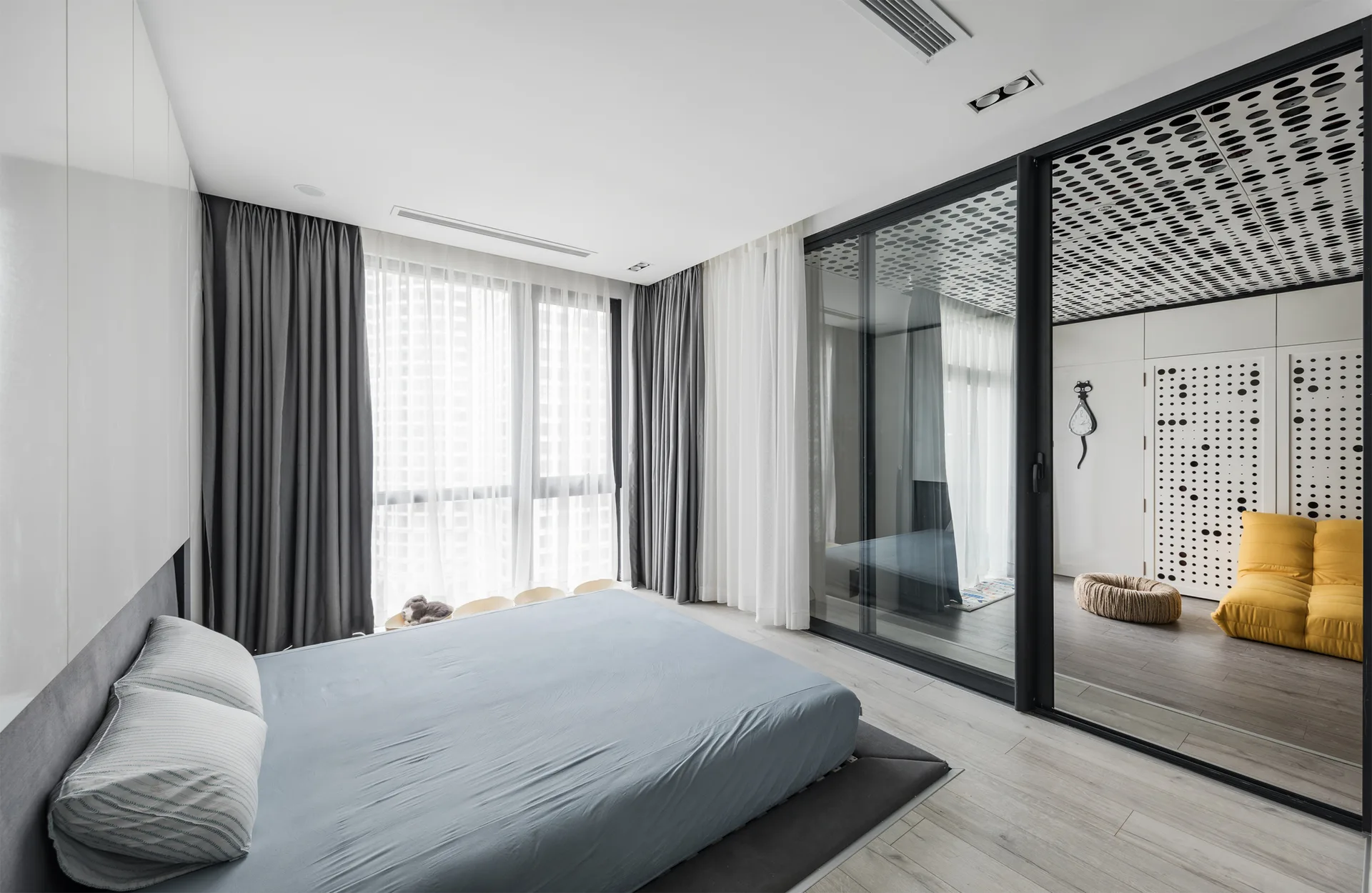 A minimalist bedroom featuring a low platform bed with blue bedding and a black-framed sliding glass partition looking into an adjacent room.