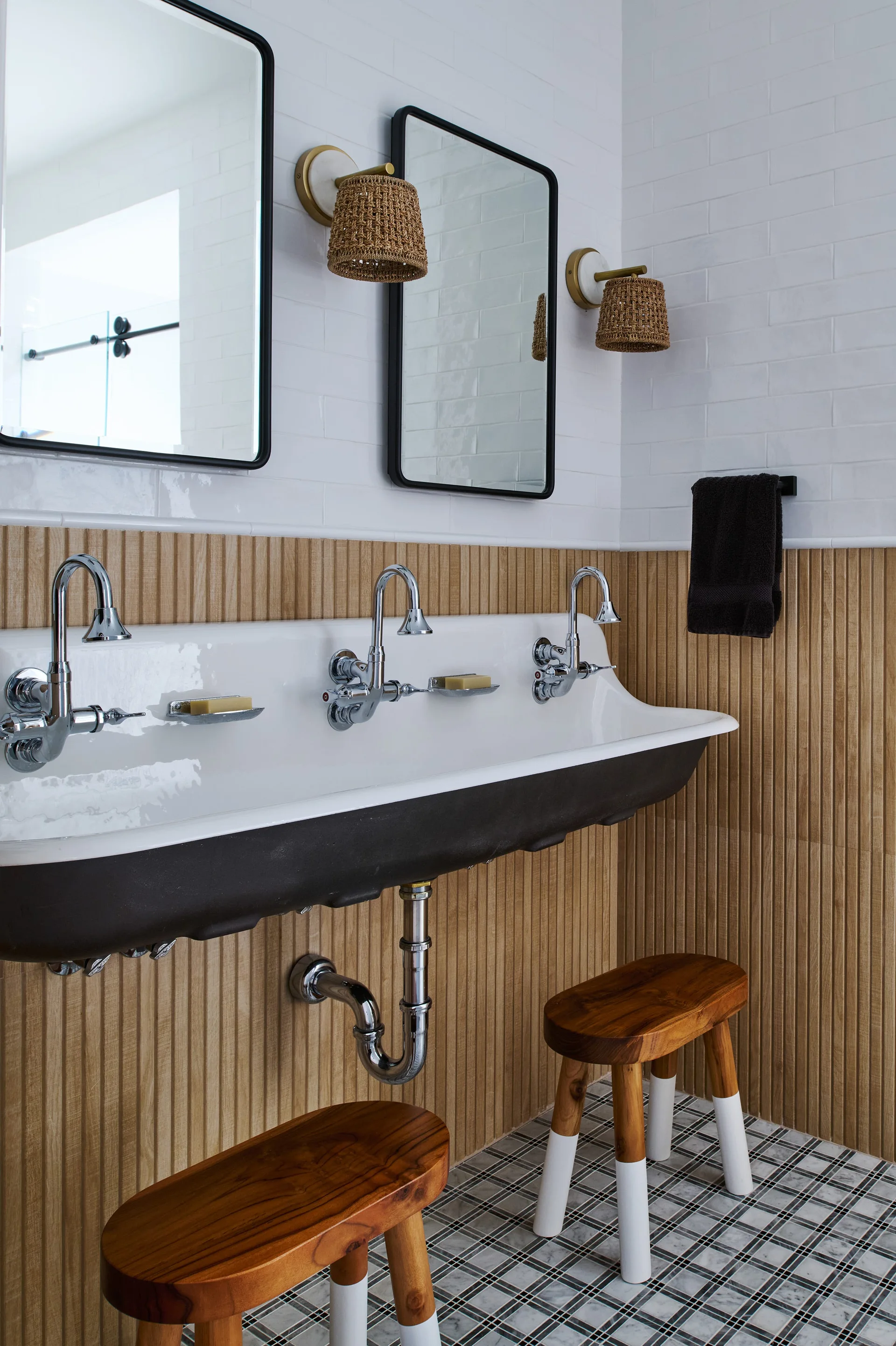 A bathroom features a wall-mounted white trough sink with three chrome faucets against a slatted wood wall under two mirrors and woven sconces.