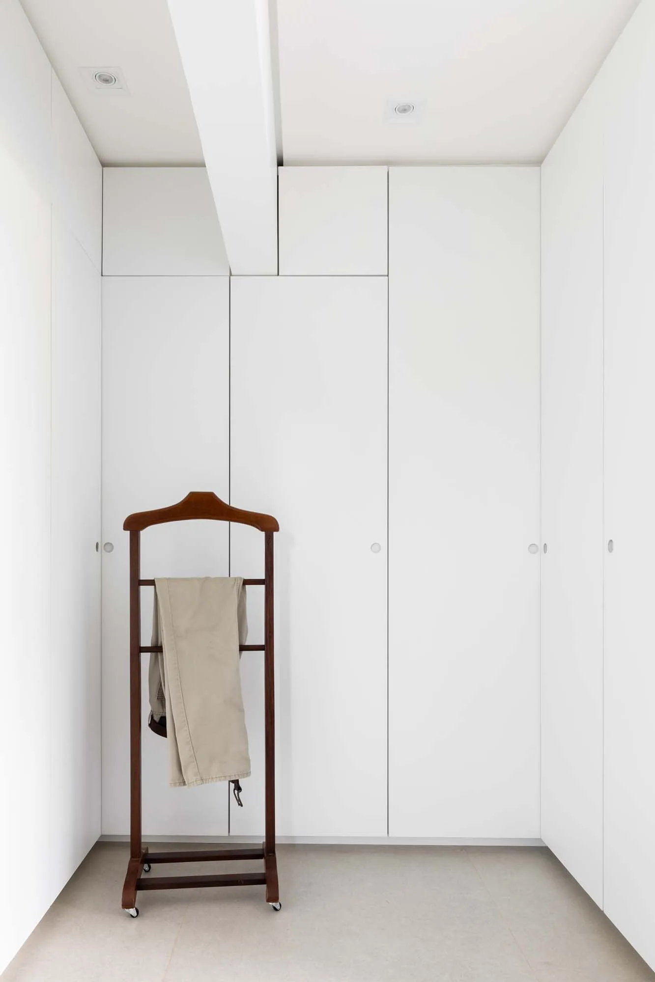 A minimalist dressing room features seamless white floor-to-ceiling wardrobes and a dark wood clothes valet stand holding folded trousers.
