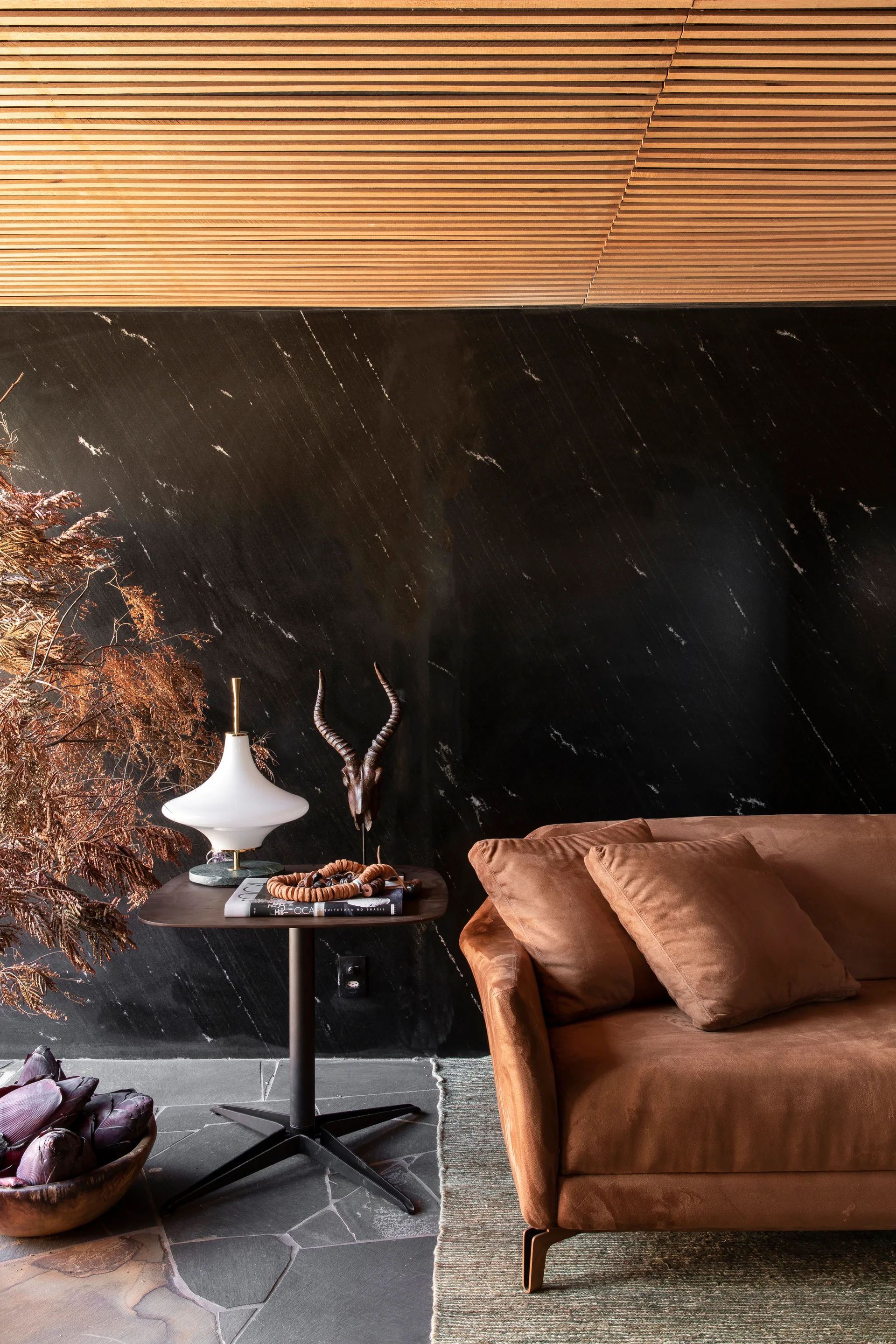 A brown fabric sofa sits on a flagstone floor against a veined black marble wall underneath a slatted wood ceiling.