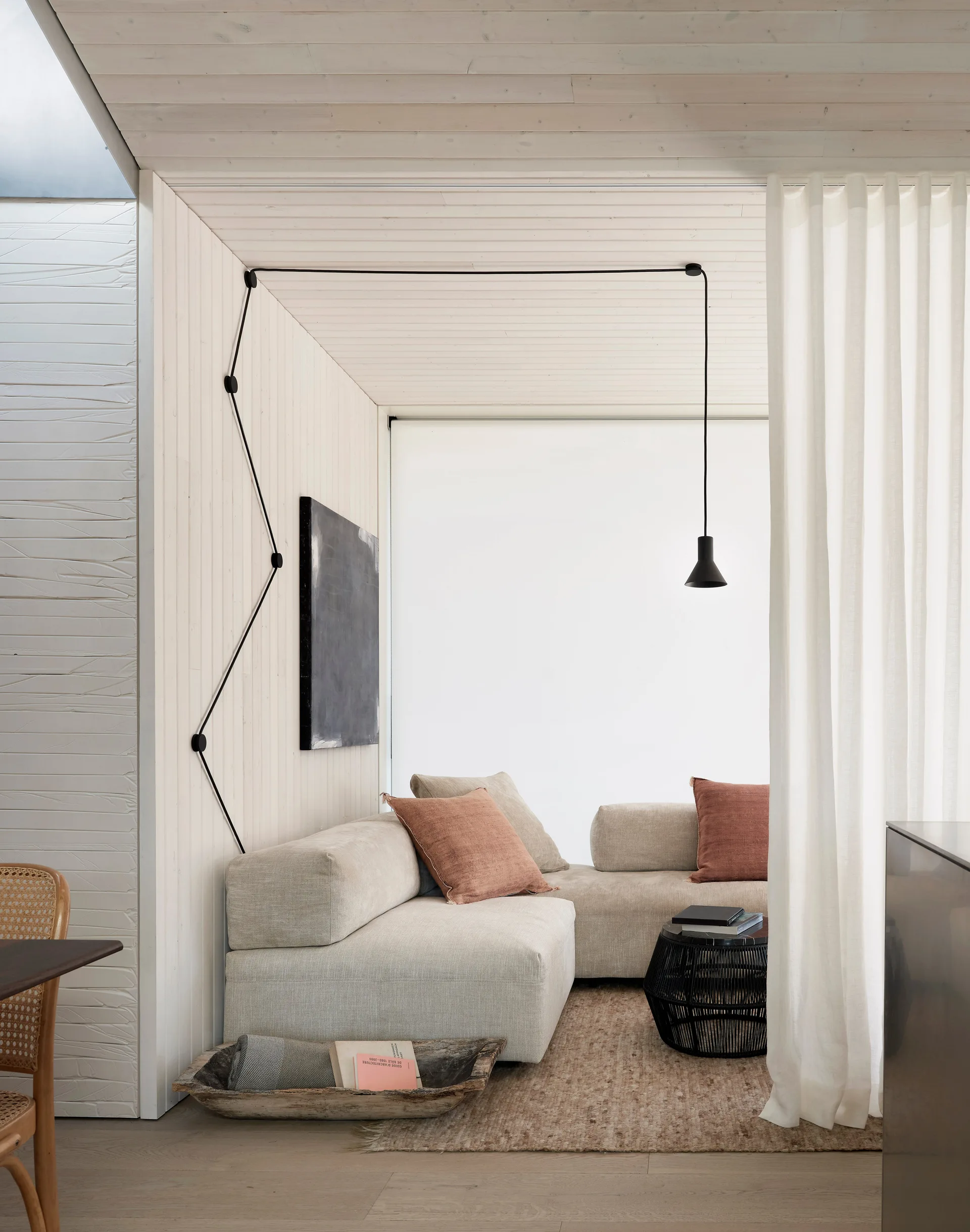 A minimalist living room corner featuring a modular beige sofa, a black pendant light with a graphic cord on white paneled walls, and a sheer curtain.