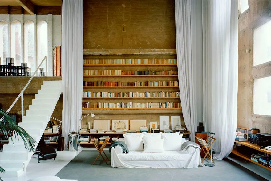 A high-ceiling industrial loft living room with a white slipcovered sofa, a built-in wooden bookcase on a concrete wall, and tall white curtains.