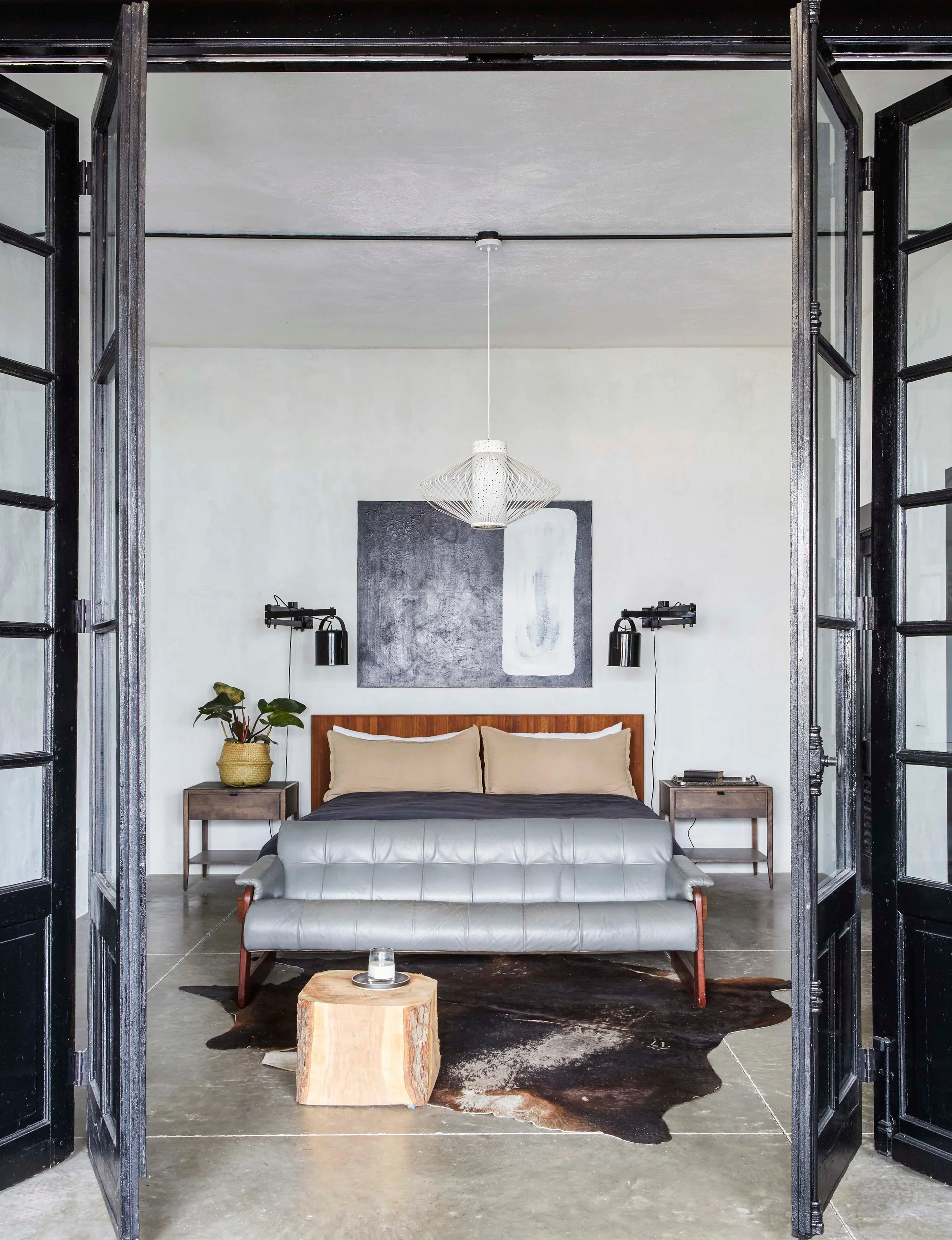 A modern bedroom seen through black-framed glass doors features a wood bed, a gray leather bench, concrete flooring, and a cowhide rug.