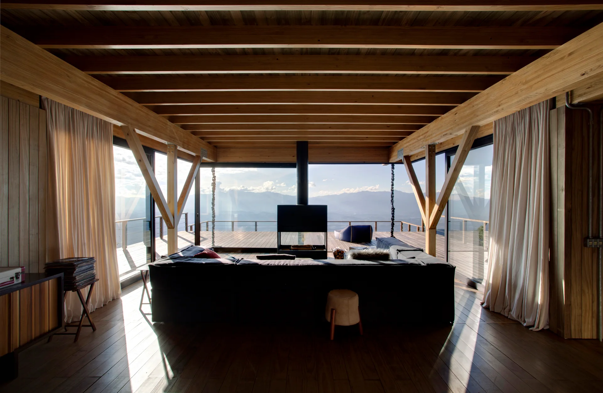 A mountain cabin living room features a dark sectional sofa, a central suspended fireplace, and exposed timber ceiling beams leading to a deck.