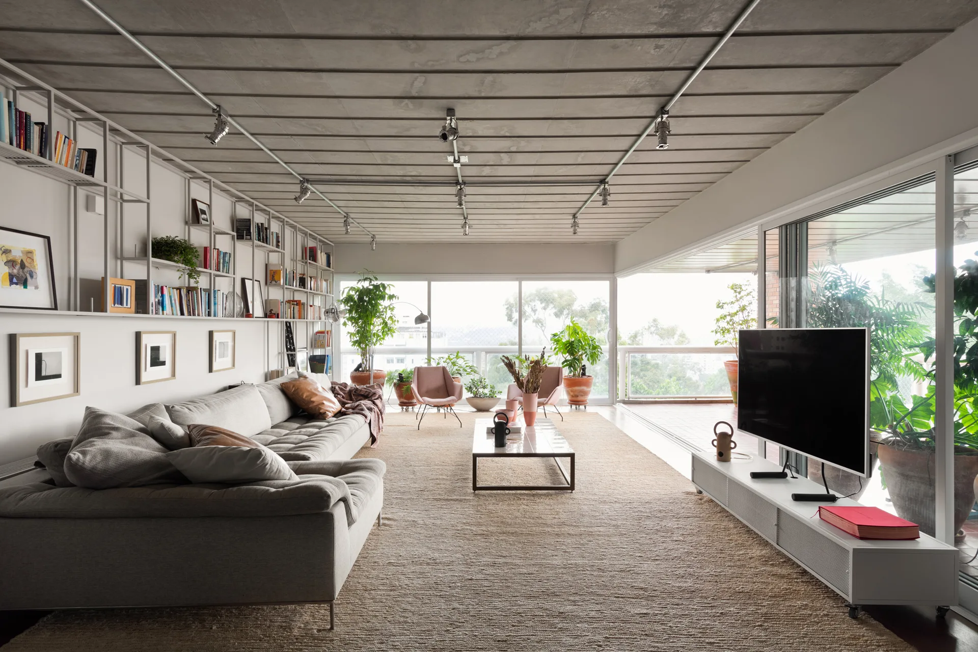 A spacious living room features a grey sectional sofa, exposed concrete ceiling with track lighting, and wall-mounted metal shelving next to balcony doors.