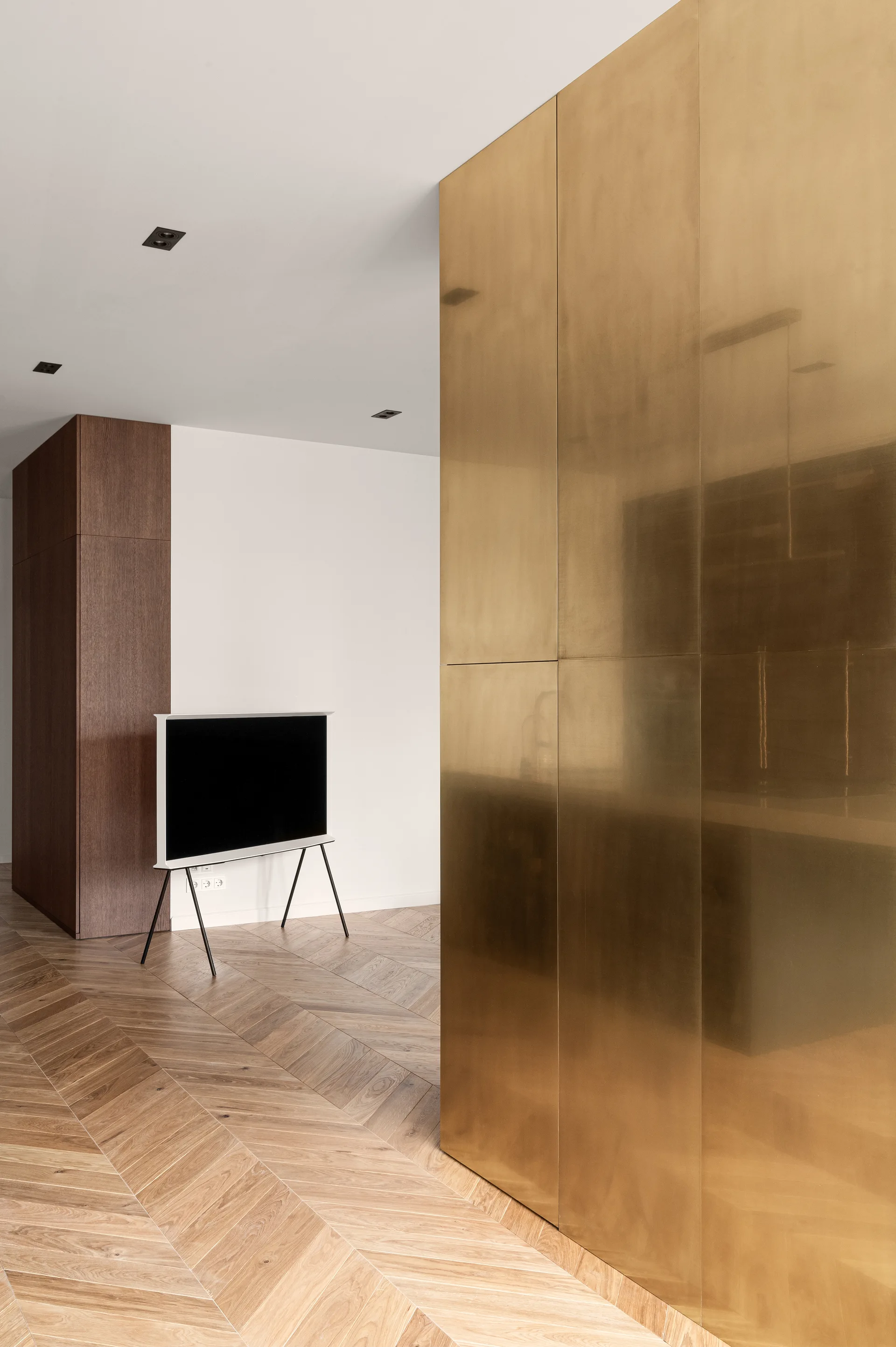 A minimalist room featuring a large brushed brass wall, a flat-screen television on a tripod stand, and herringbone pattern wood flooring.