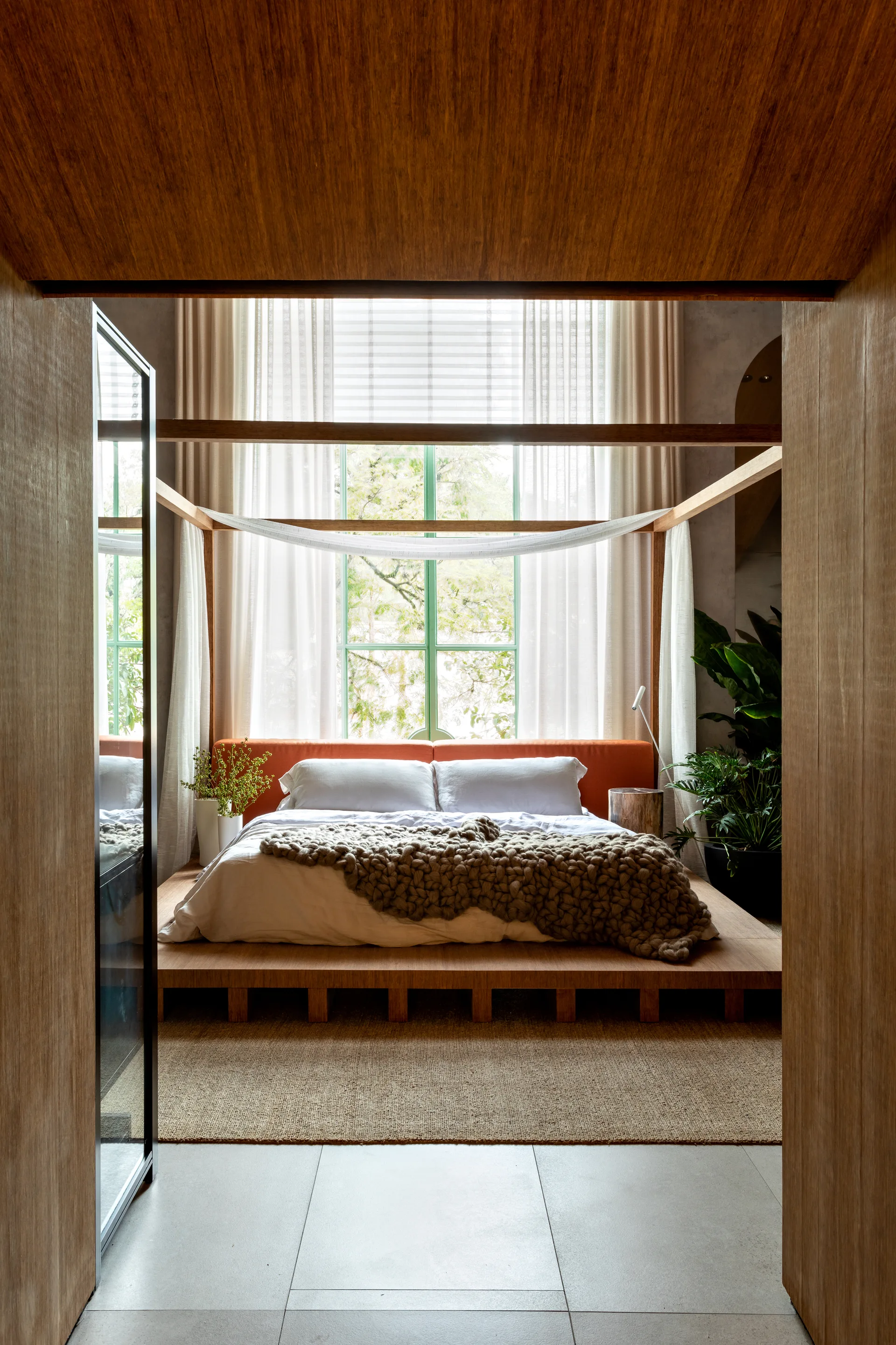 A low wooden platform bed with a matching canopy frame stands before a large window draped in sheer white curtains in a modern bedroom.