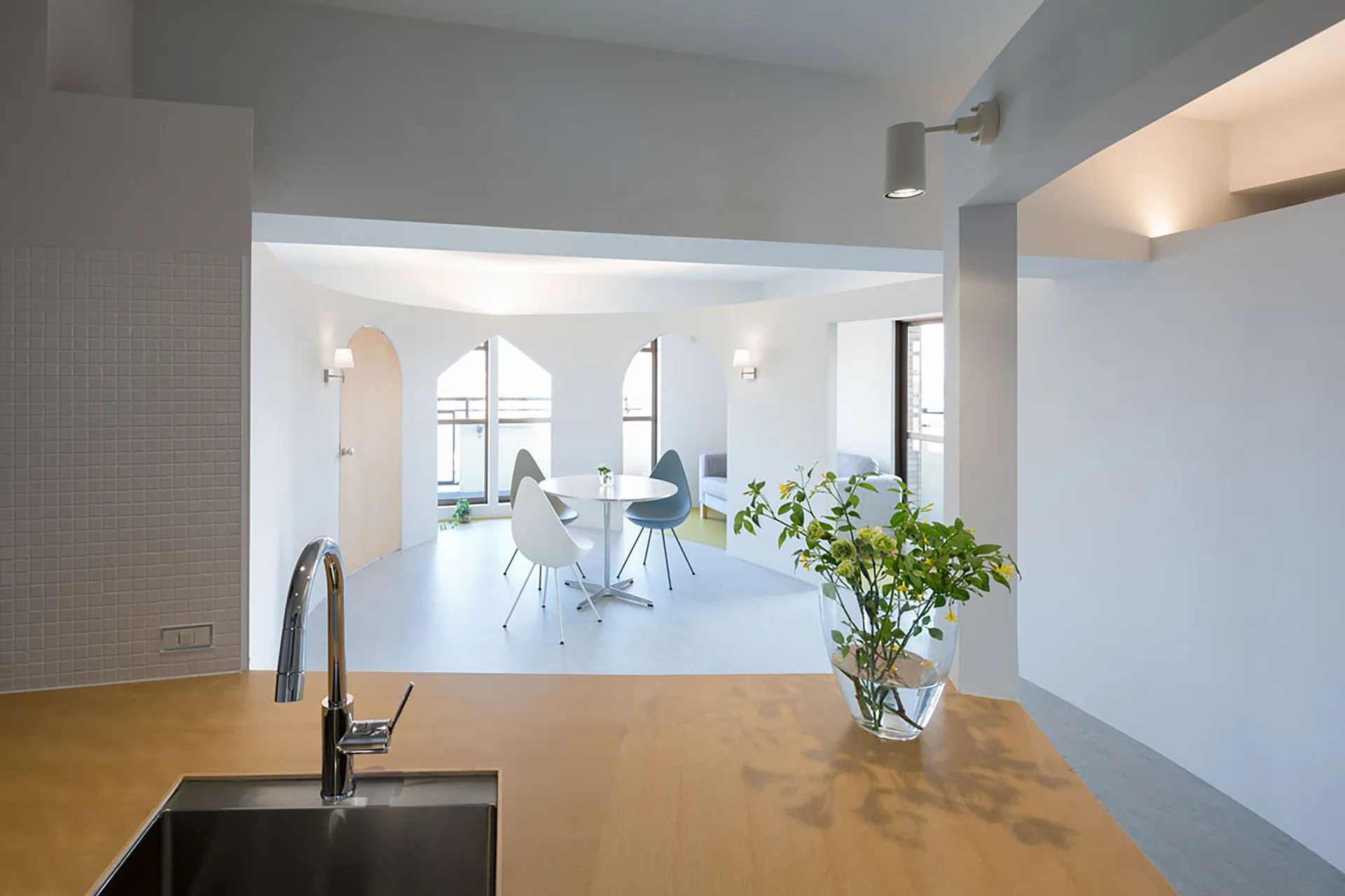A modern kitchen foregrounds a wooden island and sink, looking out toward a minimalist dining area framed by white curved walls with arched openings.