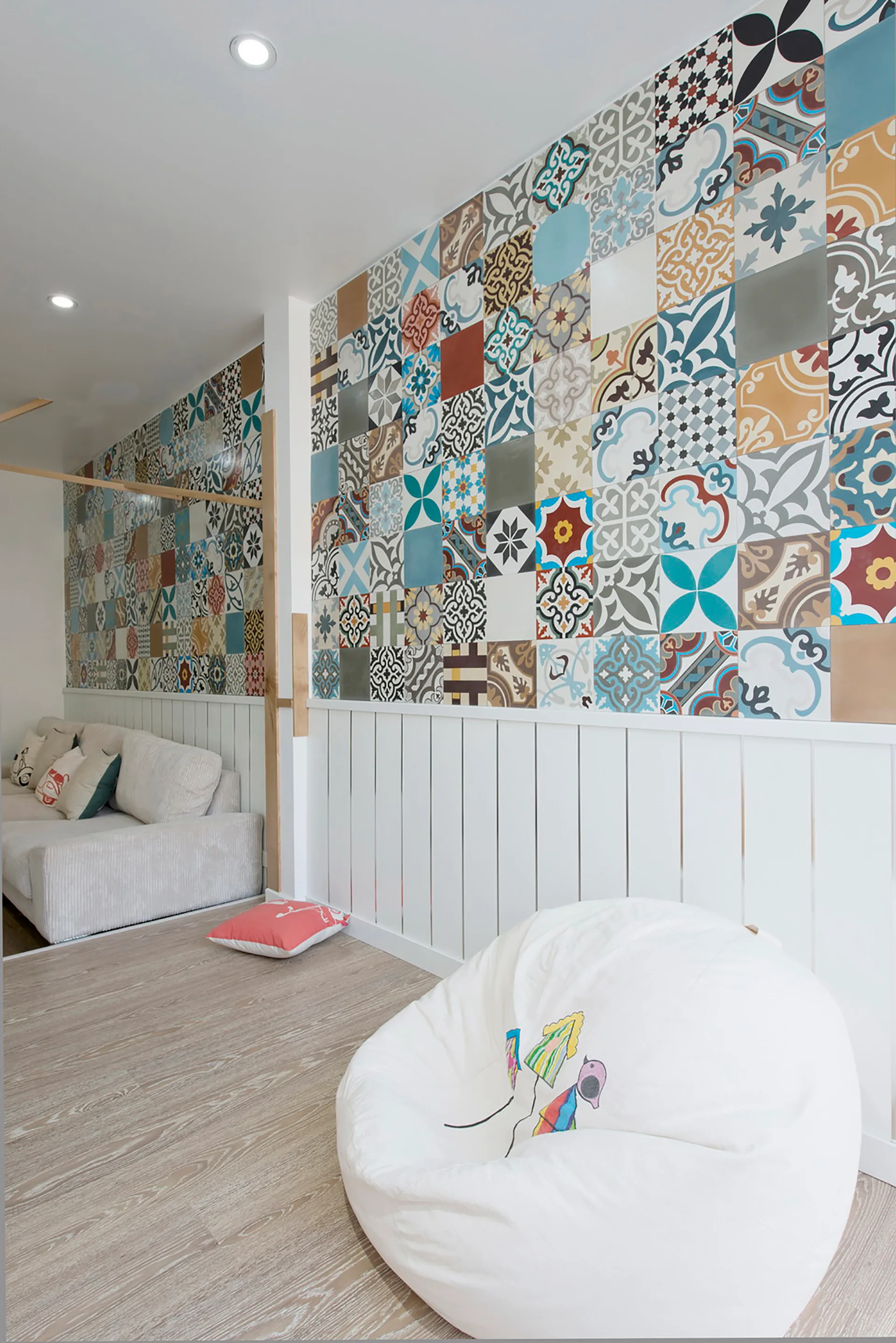 A room featuring a multicolored patterned tile accent wall above white vertical wainscoting with a white bean bag chair on a light wood floor.