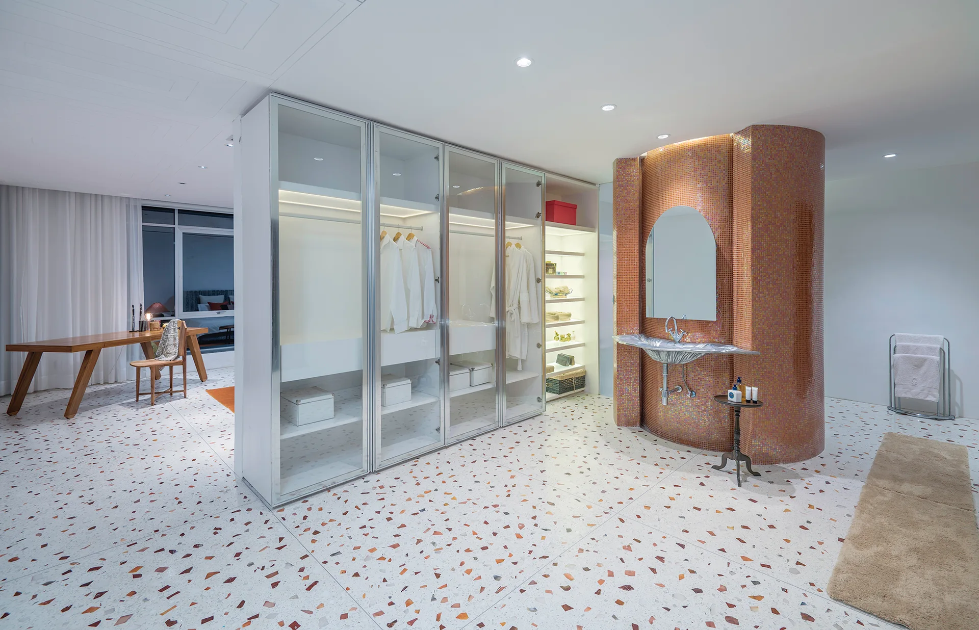 A spacious dressing room with a large glass-fronted closet system, a curved copper mosaic tile vanity pillar, and polished terrazzo flooring.