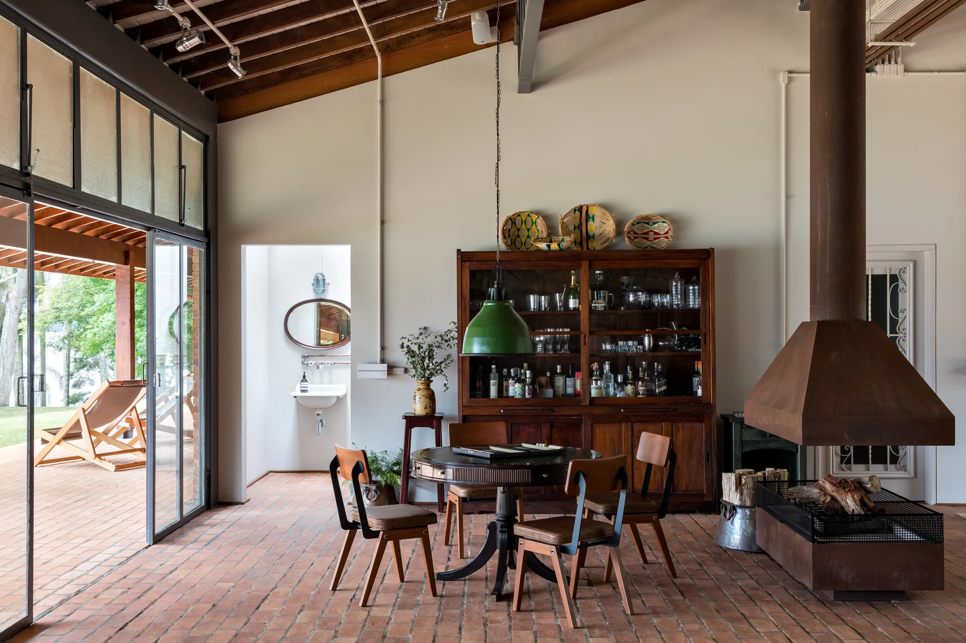 A rustic dining room features a round wooden table, four chairs, a tall display hutch, a metal fireplace hood, and red brick floors.