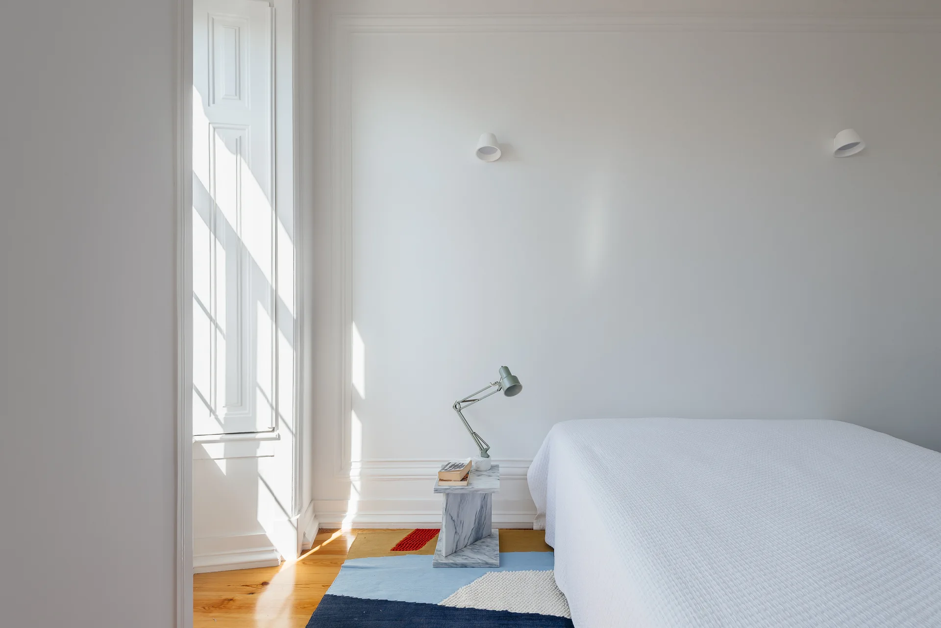 A minimalist bedroom features a white bed next to an angular marble side table, a green task lamp, and a multi-colored geometric rug.