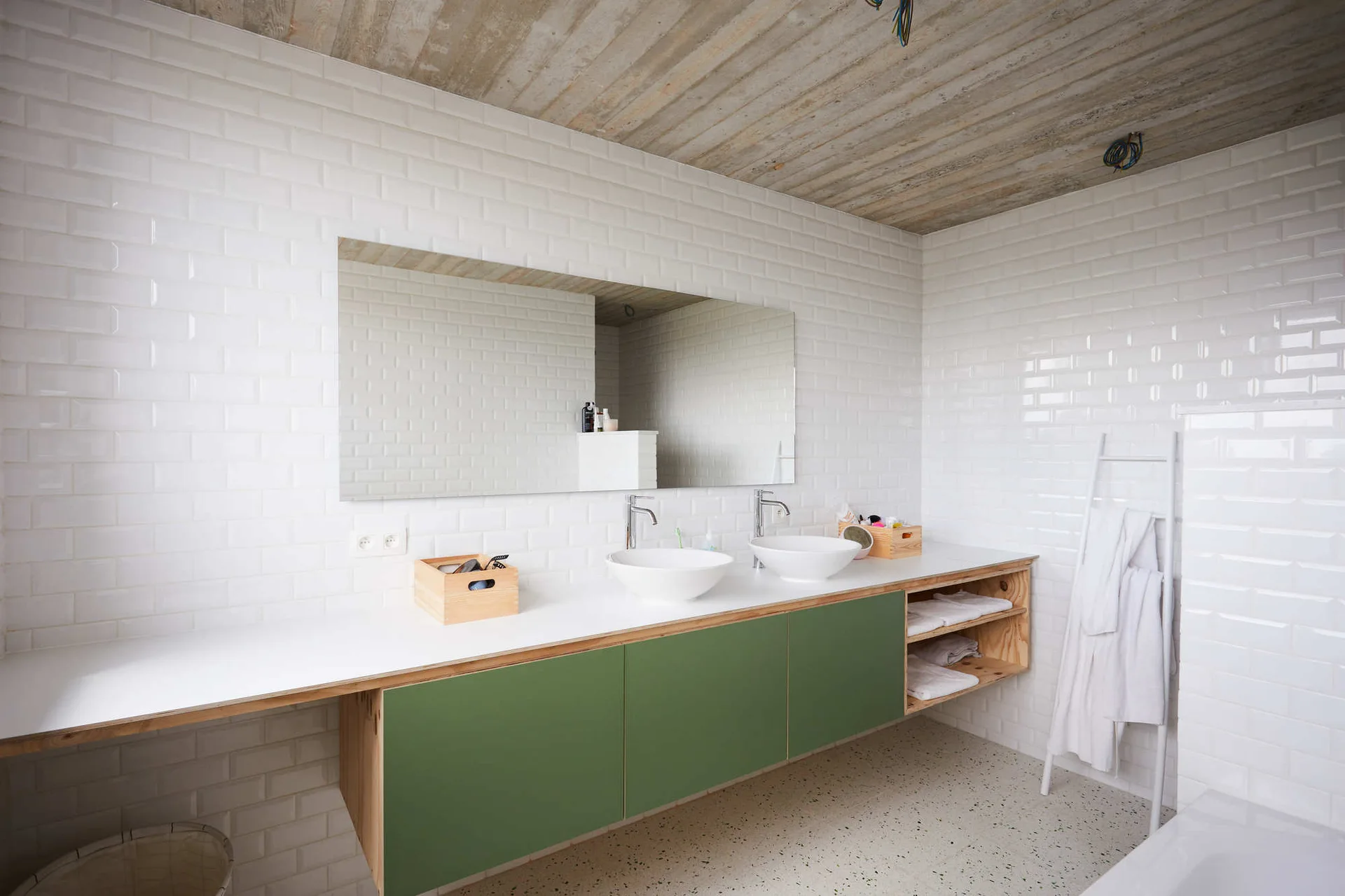 A contemporary bathroom features a long green and wood floating vanity, white subway tile walls, dual vessel sinks, and an exposed concrete ceiling.