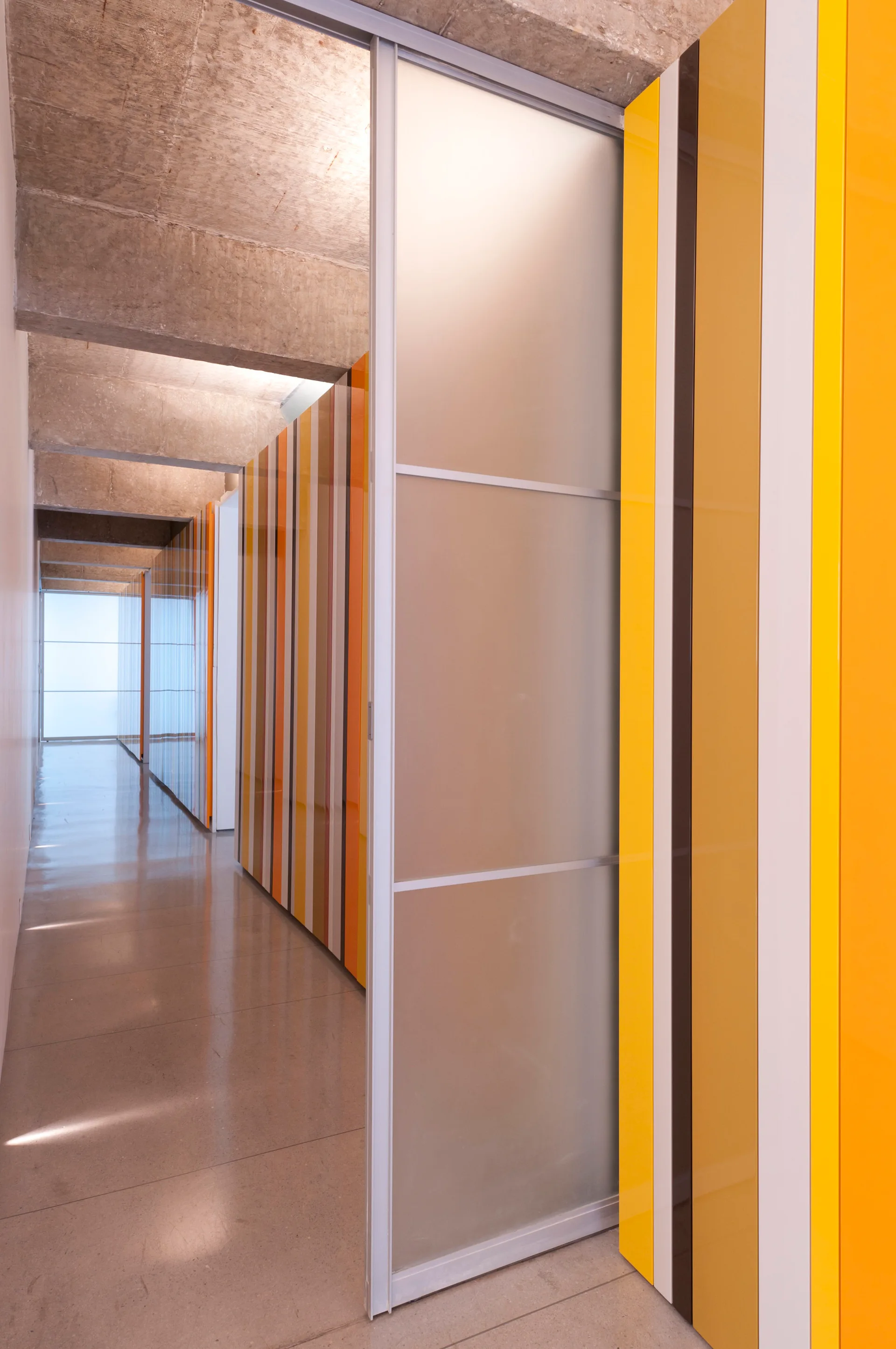A modern hallway featuring polished concrete floors, exposed ceiling beams, a frosted glass sliding door, and glossy orange and yellow striped wall panels.